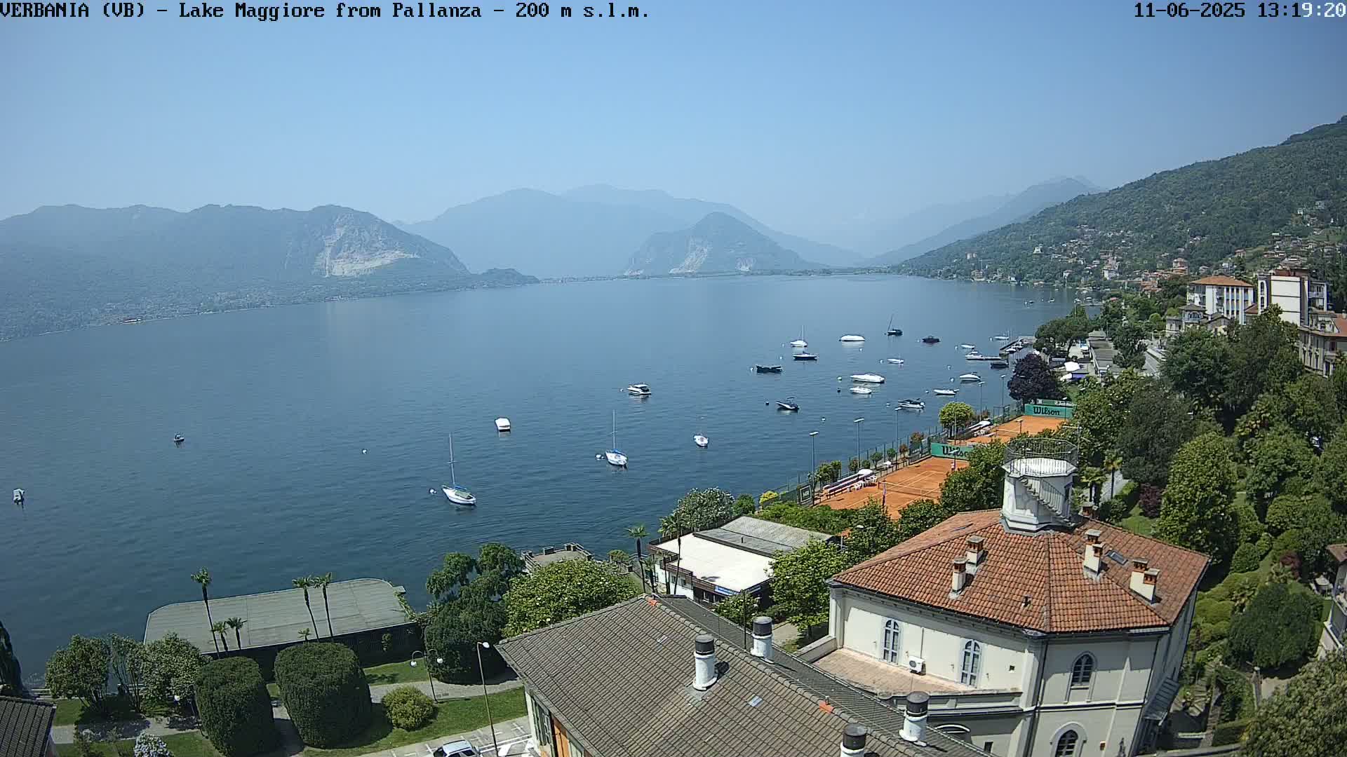 A clear, sunny day reveals a calm lake with numerous boats, tennis courts, and buildings nestled among lush greenery, all set against a backdrop of mountains.