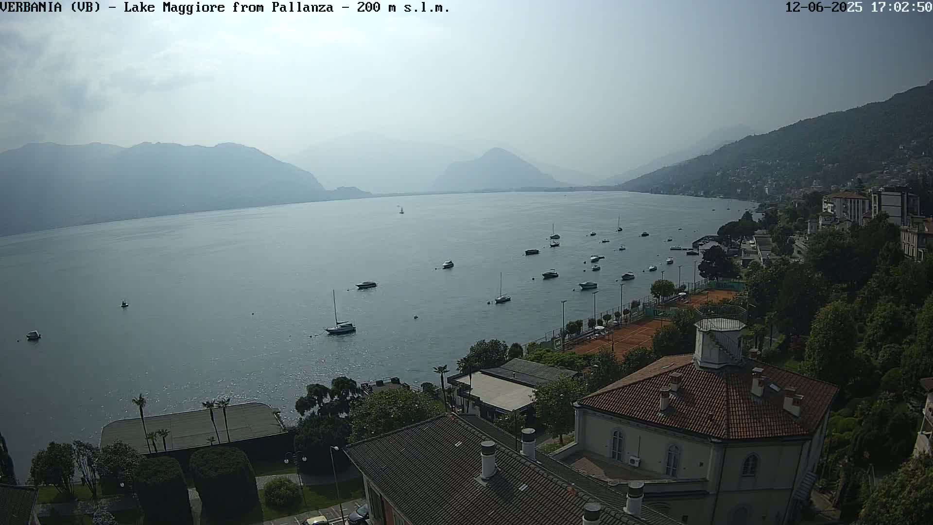 A hazy, sunny day overlooks a calm lake with numerous boats, tennis courts, and buildings nestled along a hillside.