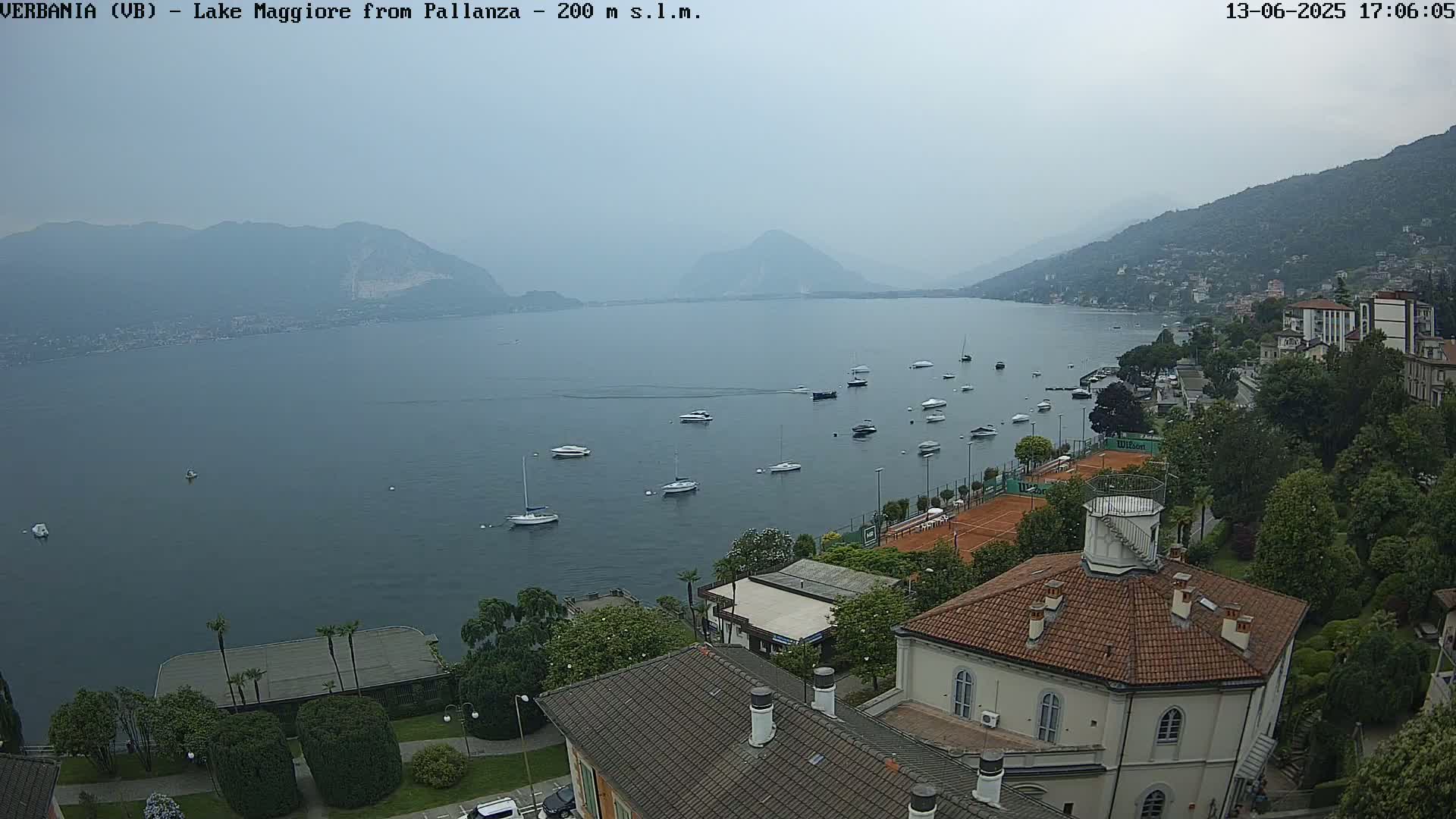 A hazy view of a lake with numerous boats, surrounded by hills, buildings, and tennis courts.