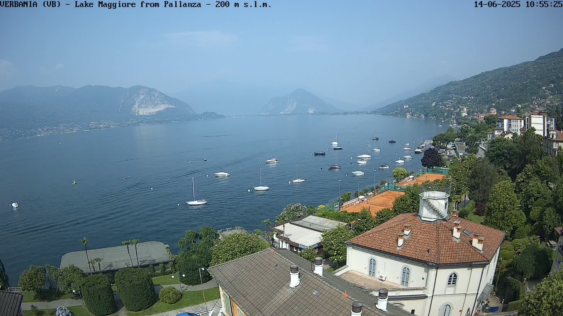 A high-angle view on a sunny day shows Lake Maggiore with numerous boats, a tennis court, and buildings along the shoreline, nestled between mountains.