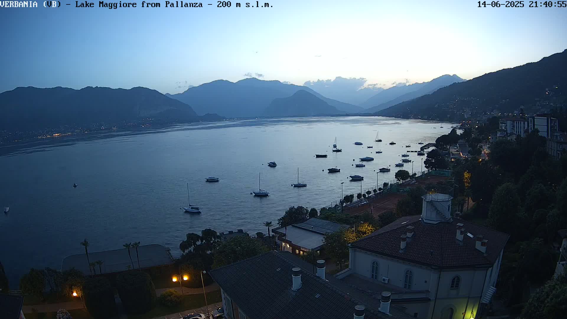 A view of Lake Maggiore from a high vantage point on an overcast day, showing several boats on the water, tennis courts, and buildings nestled along the shoreline.