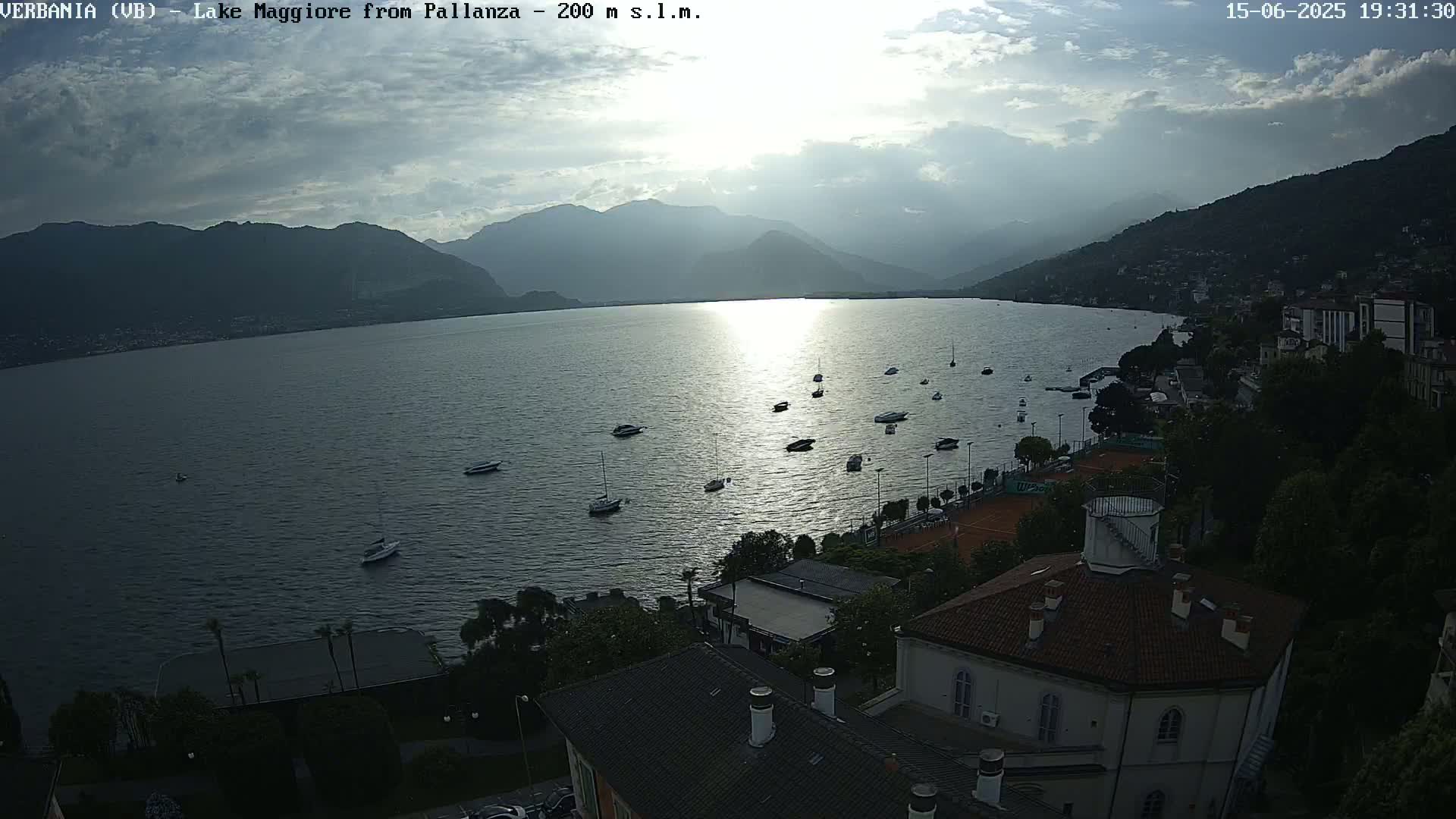 A partly cloudy sunset over a lake with numerous boats, seen from an elevated vantage point overlooking a town nestled against mountains.