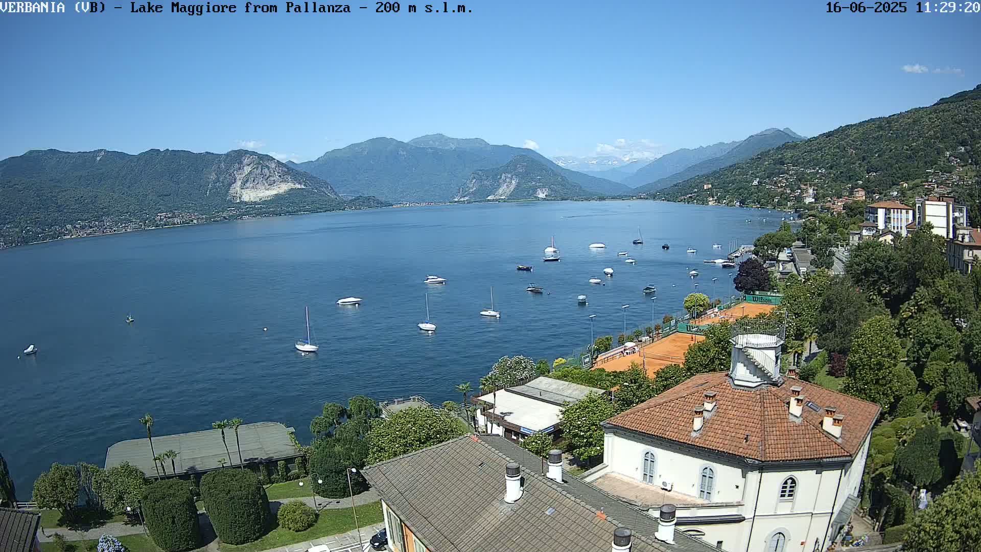 A sunny view of Lake Maggiore, surrounded by mountains and featuring several boats on the water, tennis courts, and buildings along the shoreline.