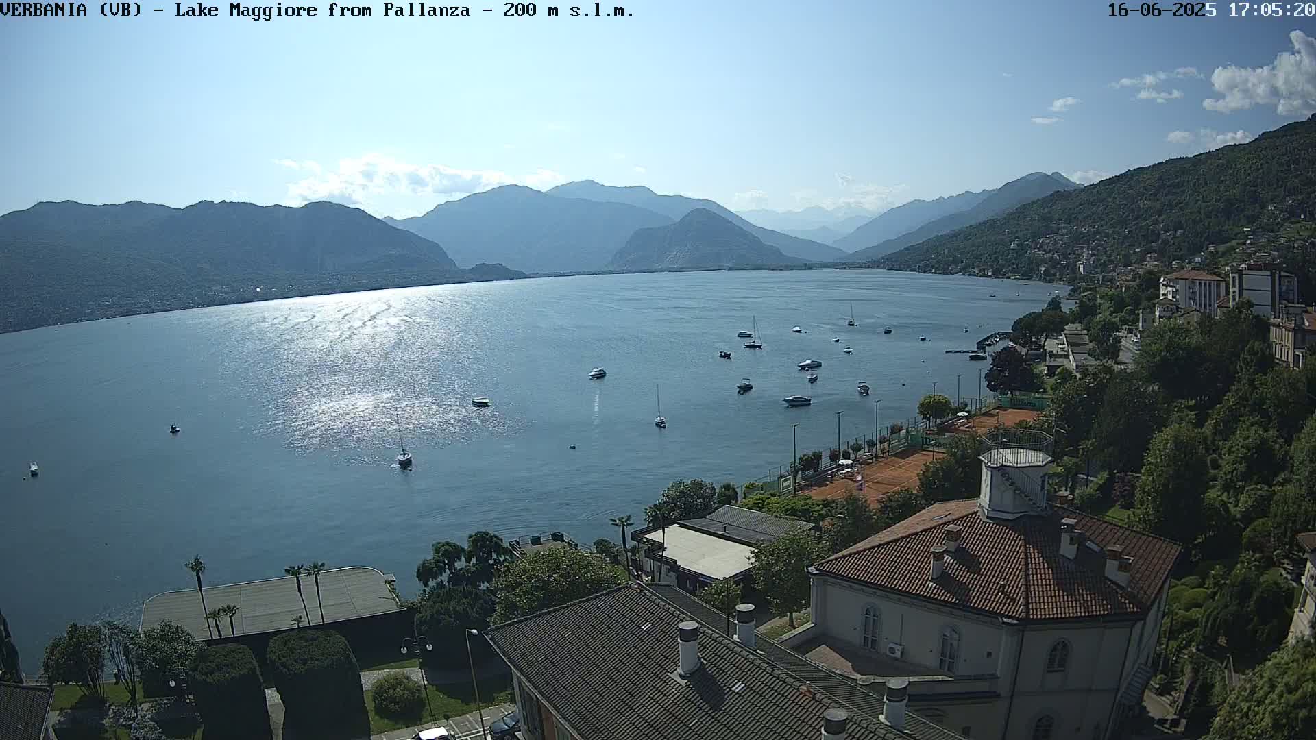 A sunny, high-angle view shows a large lake with several boats, surrounded by mountains and a lakeside town with tennis courts and buildings, including a round building with a terracotta roof.