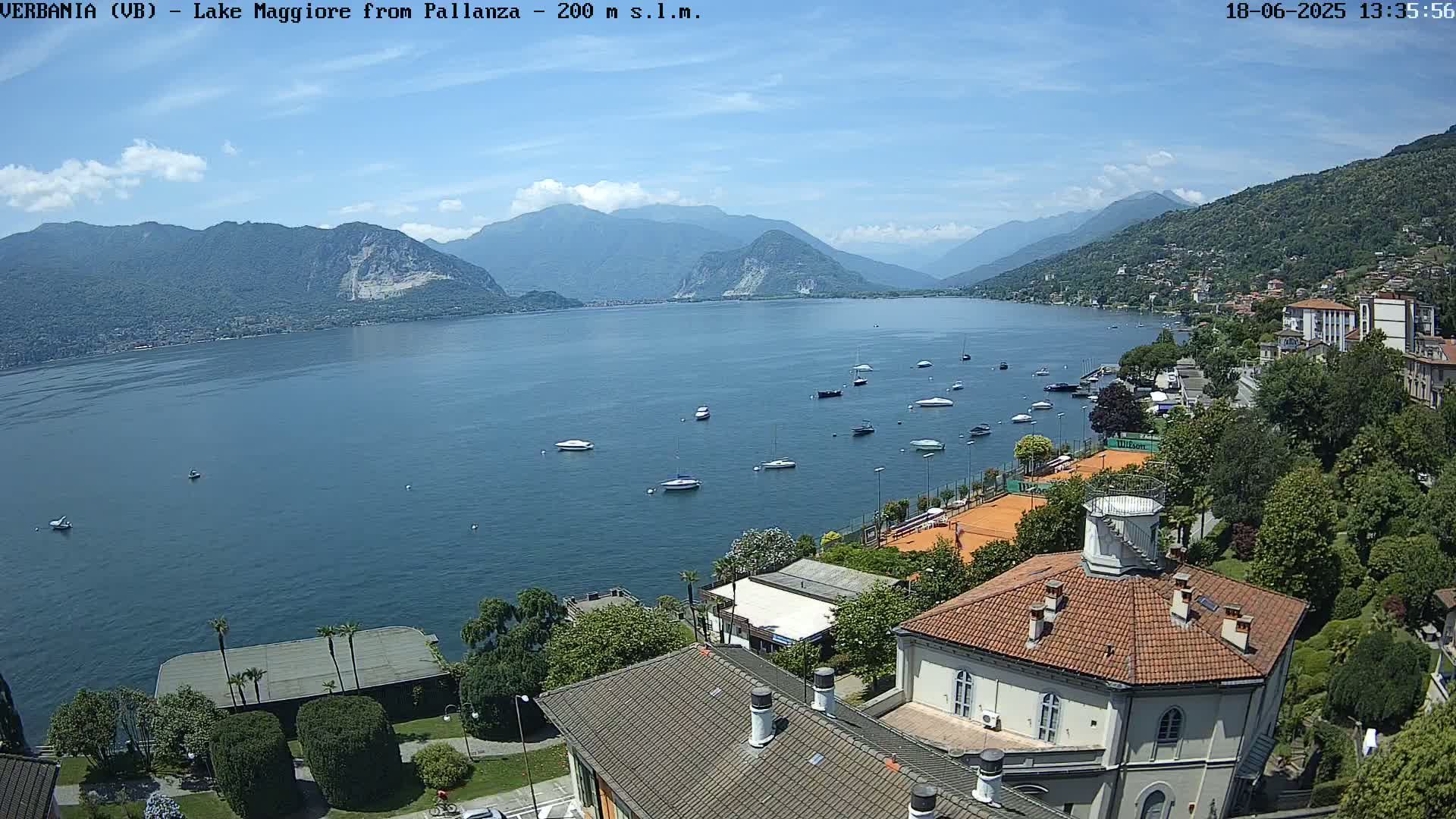 A sunny day overlooking Lake Maggiore, with numerous boats scattered on the water and a village nestled along the shoreline, surrounded by mountains.