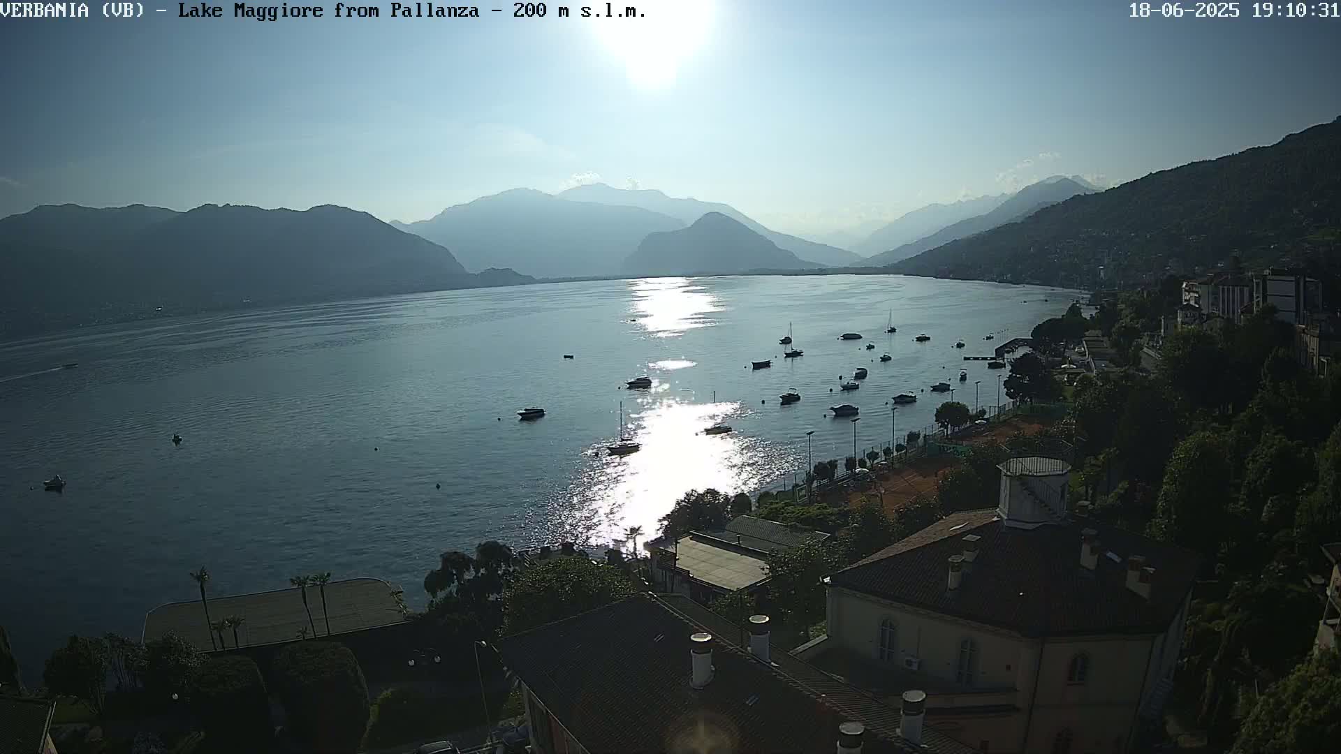 A bright, sunny view overlooking a calm lake with numerous boats, a shoreline town, and mountains in the distance.