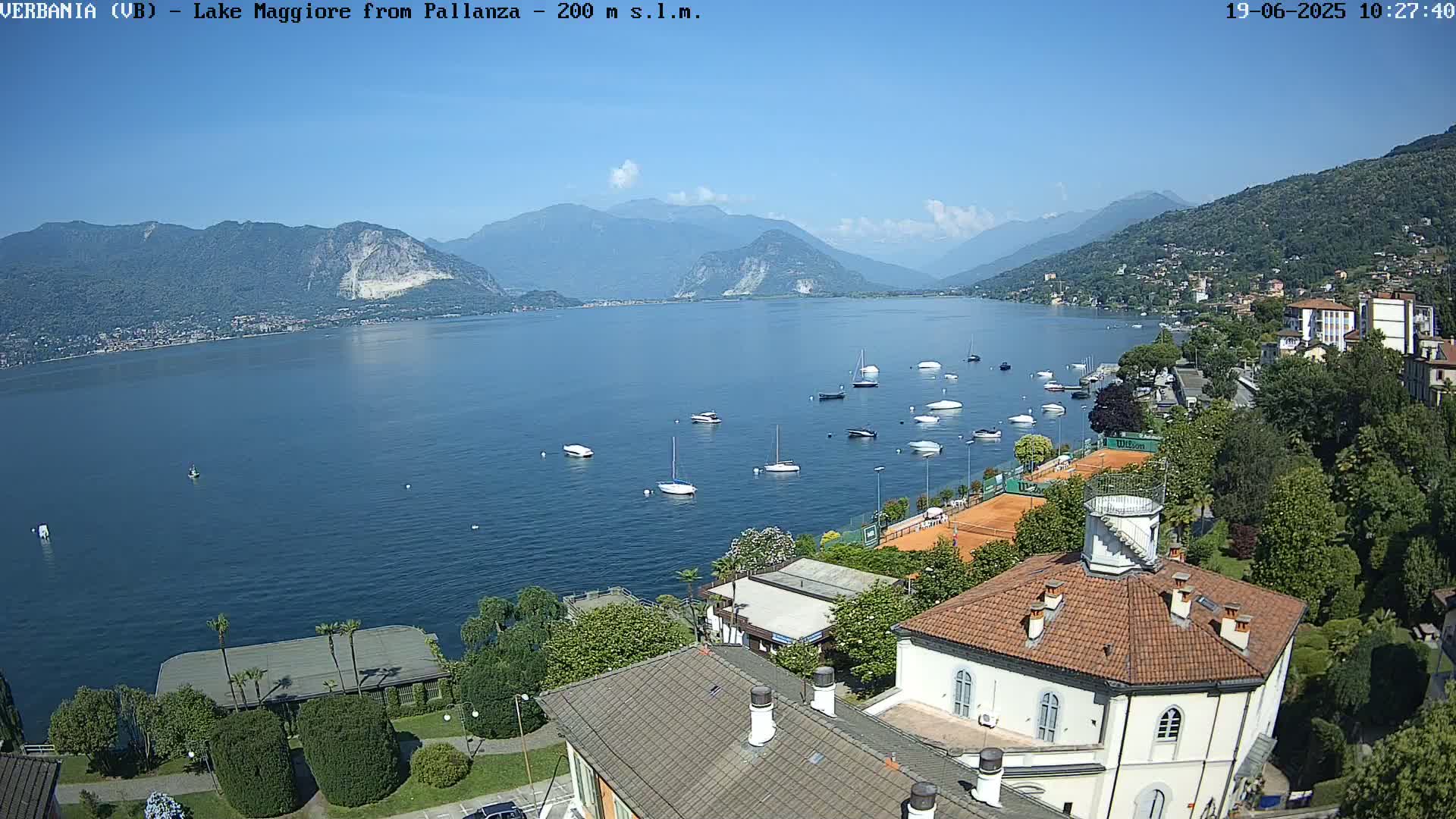 A high-angle view shows a calm lake with several sailboats, a tennis court, and buildings on a sunny day, with mountains in the background.