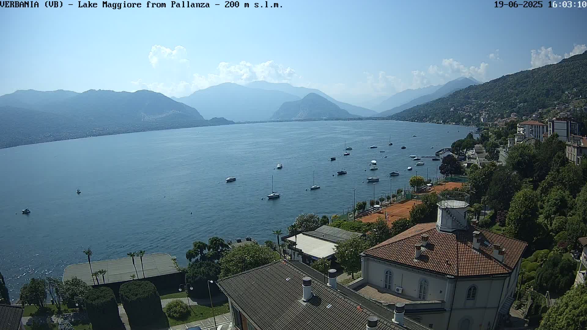 A sunny day overlooking a calm lake with numerous boats, surrounded by mountains and buildings, including a round building with a red tile roof.