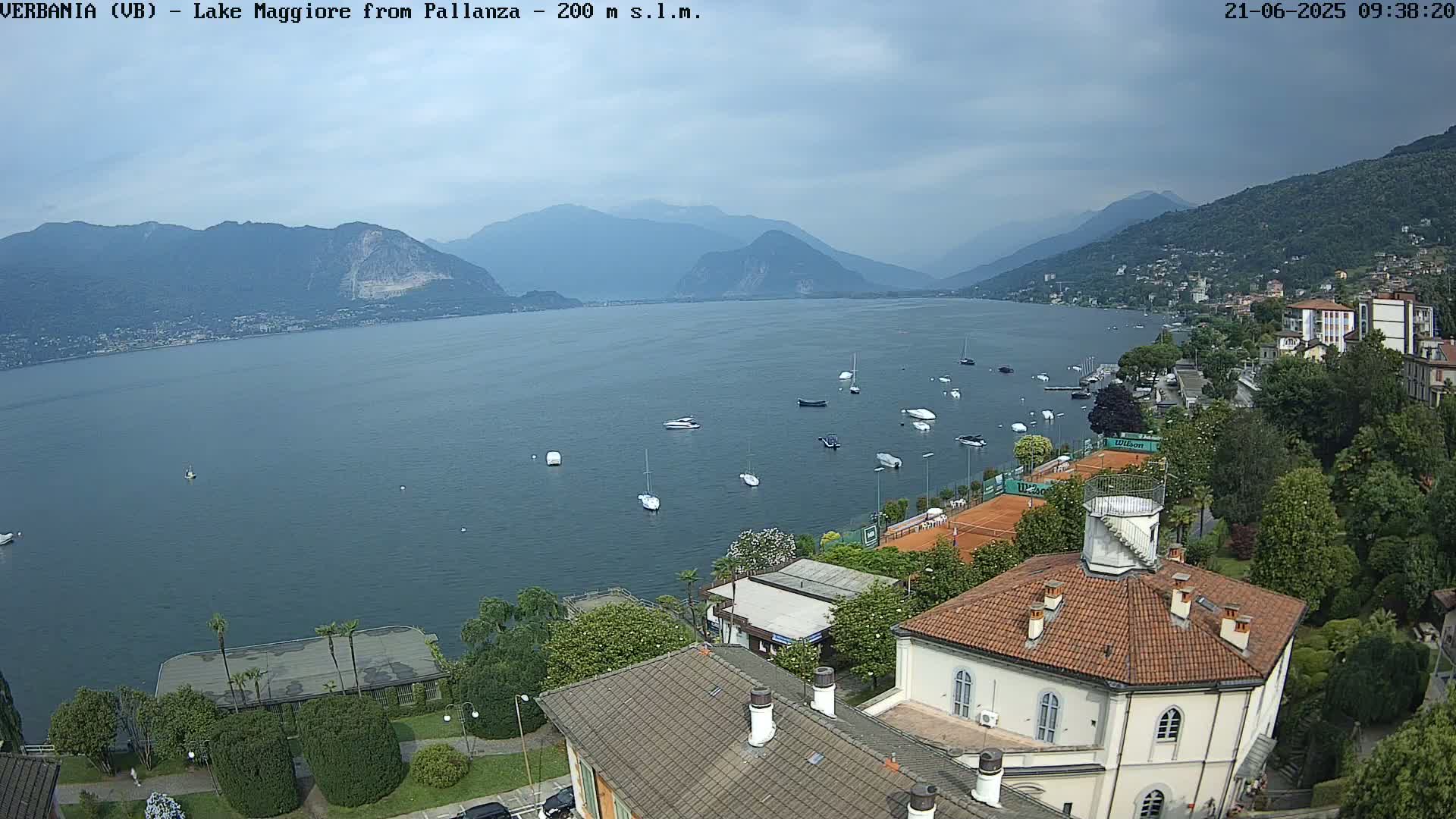 A view of Lake Maggiore from a high vantage point on an overcast day, showing numerous boats on the water, tennis courts, and buildings nestled along the shoreline with mountains in the background.