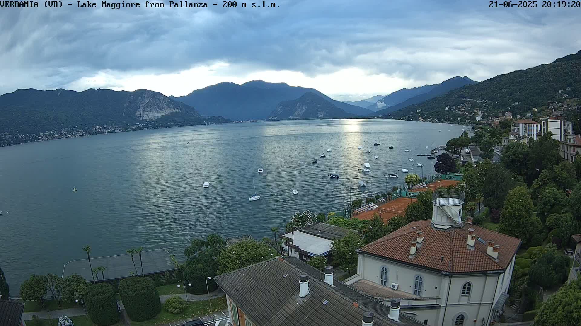 An overcast day shows a calm lake with numerous boats scattered across its surface, surrounded by mountains and buildings, including a tennis court and a building with a red-tiled roof.