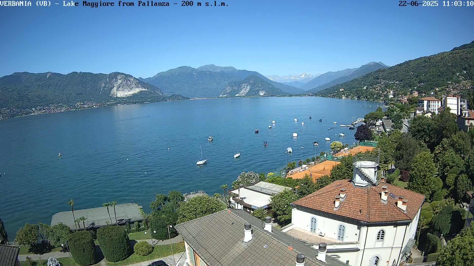 A clear, sunny day overlooking a calm lake with numerous small boats, tennis courts, and buildings nestled among lush greenery and mountains in the distance.
