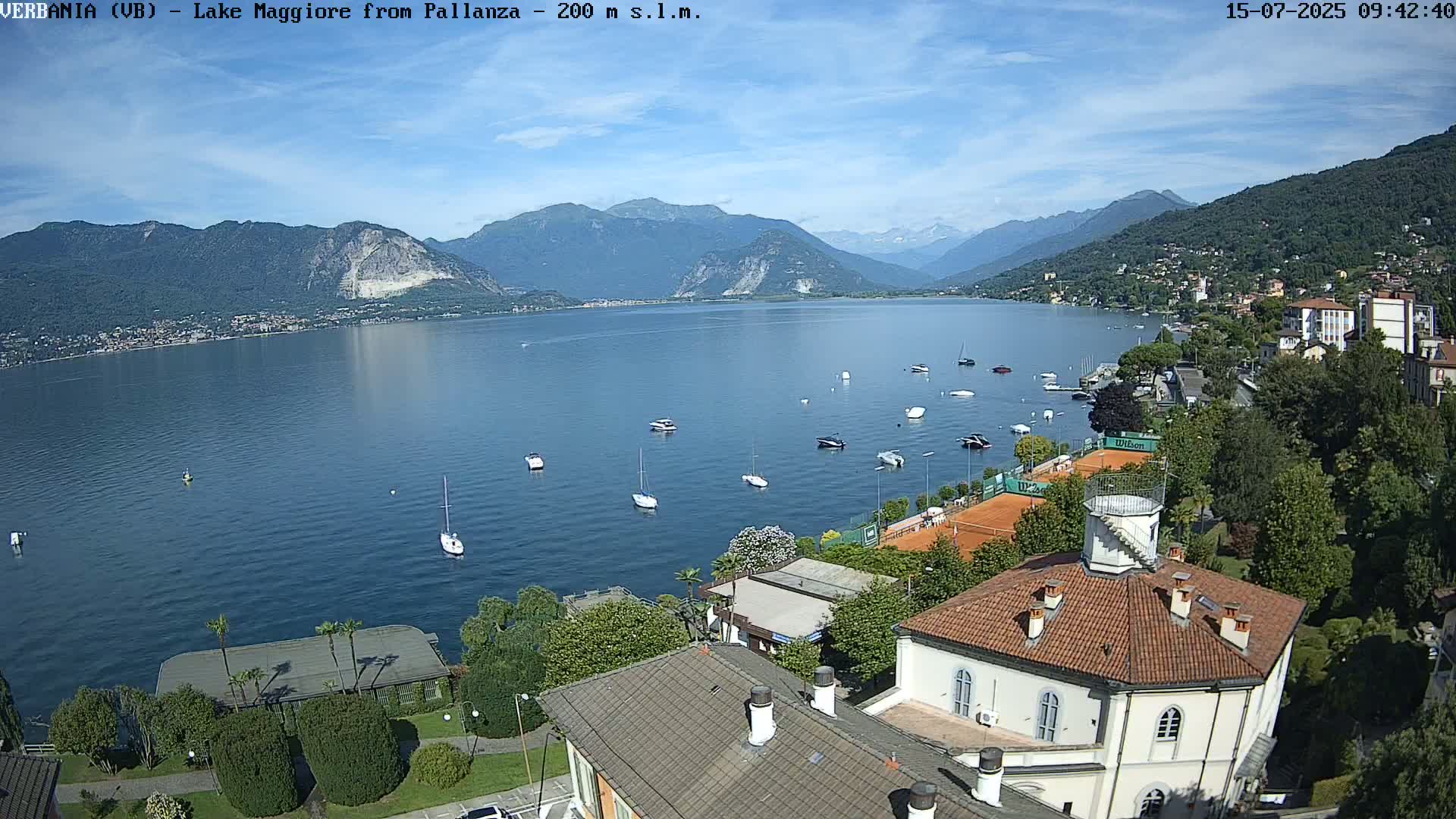 A sunny day overlooking a calm lake with numerous boats, tennis courts, and buildings nestled amongst lush greenery and mountains in the background.