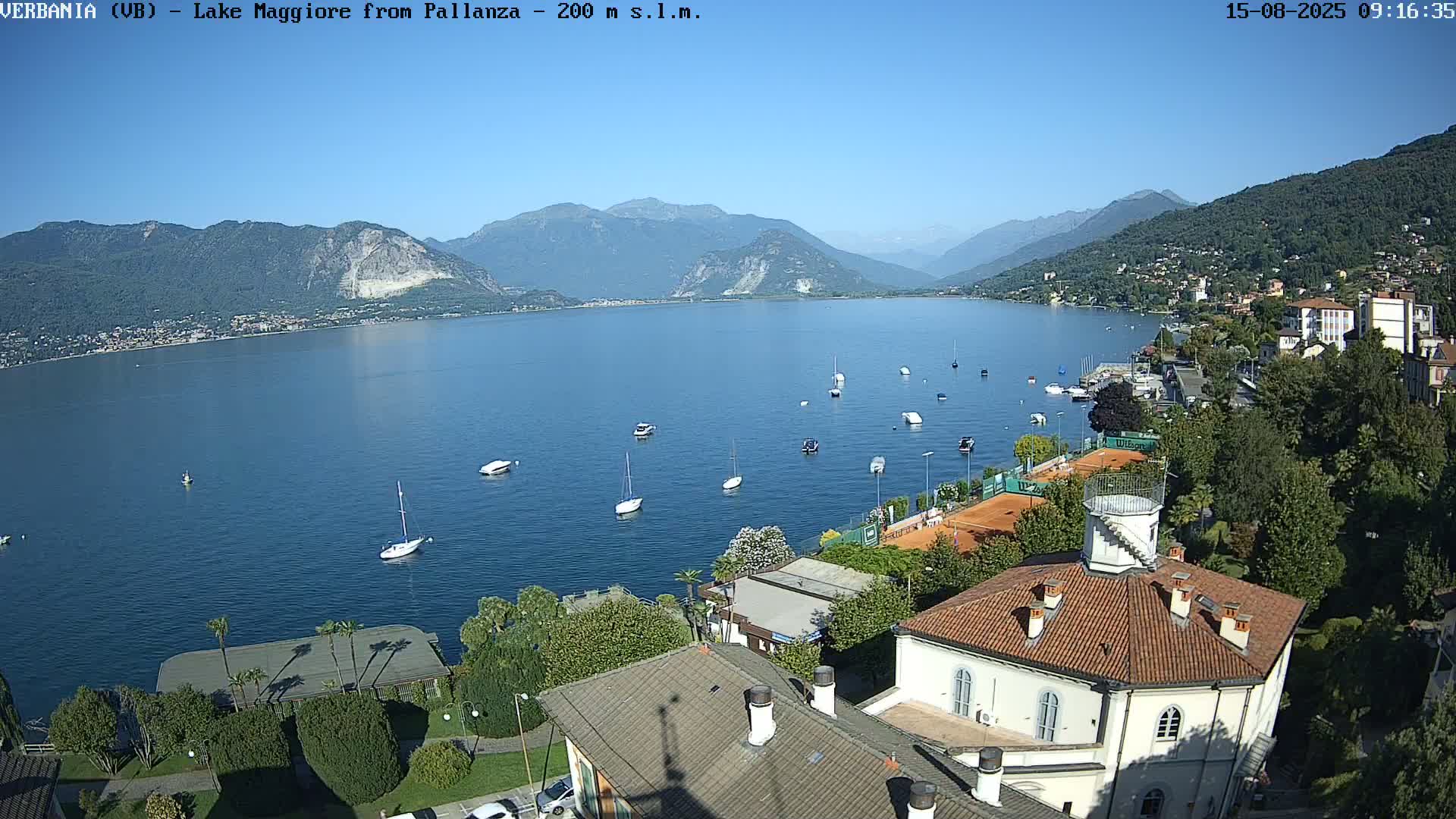 A sunny, clear day overlooks a calm lake with several boats, a tennis court, and a building with a red roof.