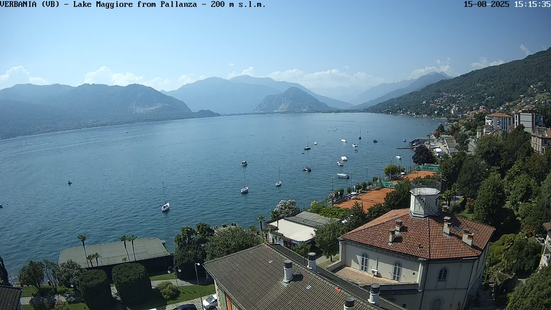 A sunny, clear day overlooks a calm lake with several boats, a tennis court, and a building with a red roof.
