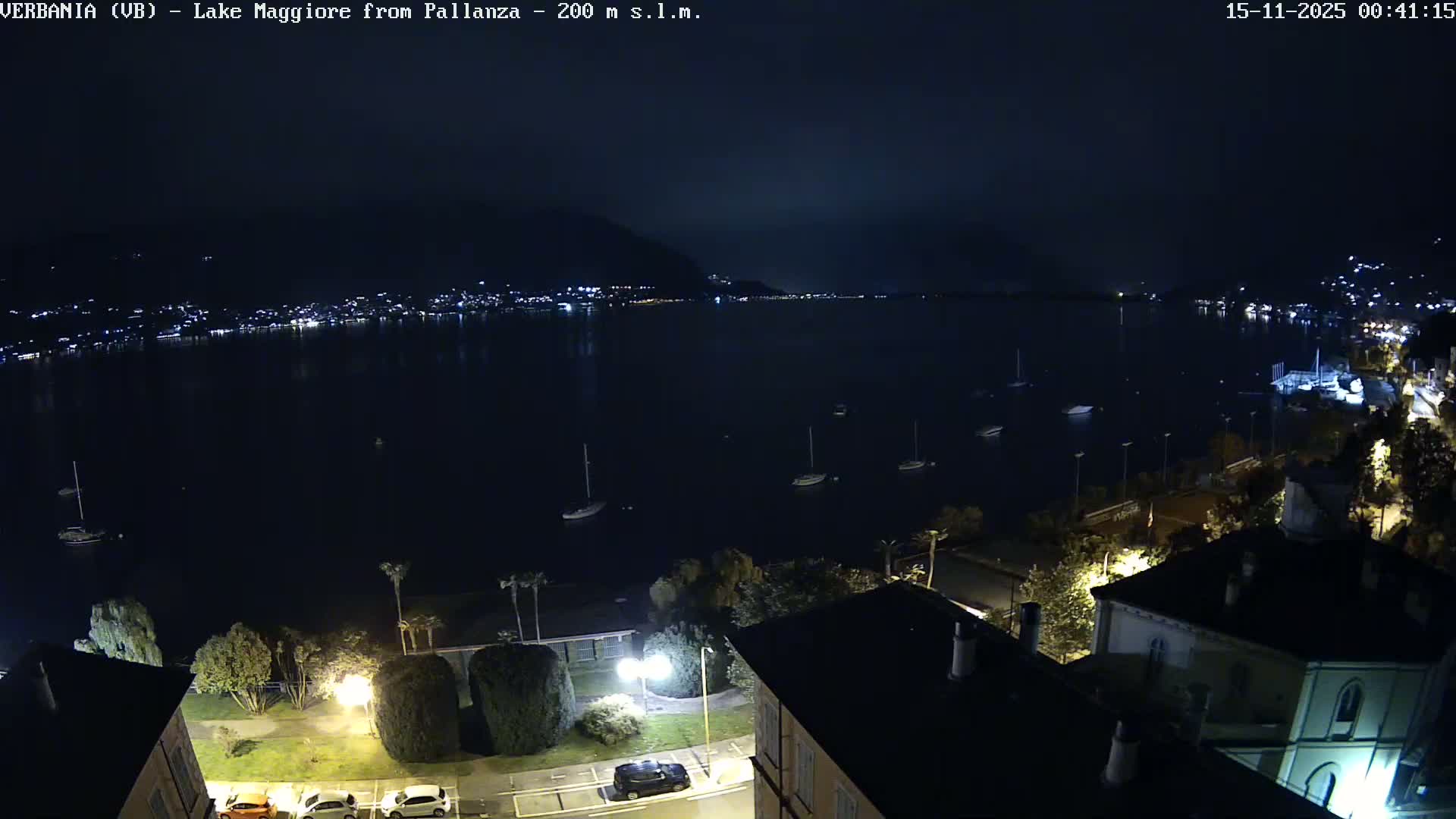 A dark, clear night view overlooks a wide body of water dotted with anchored sailboats, while the distant shoreline sparkles with city lights reflecting on the surface.