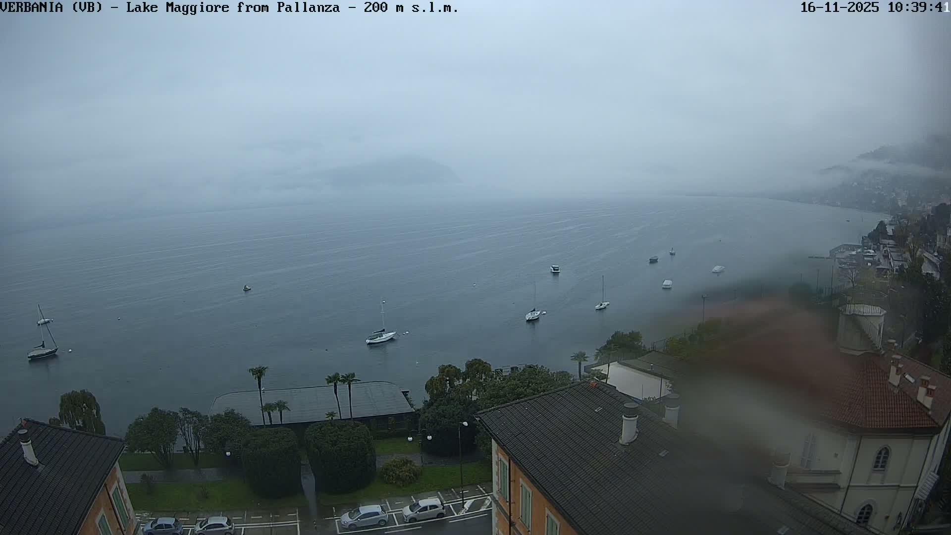 An expansive lake, dotted with numerous boats and bordered by residential buildings and green trees along its shores, appears under an overcast and hazy sky, indicating a gloomy, possibly rainy day.