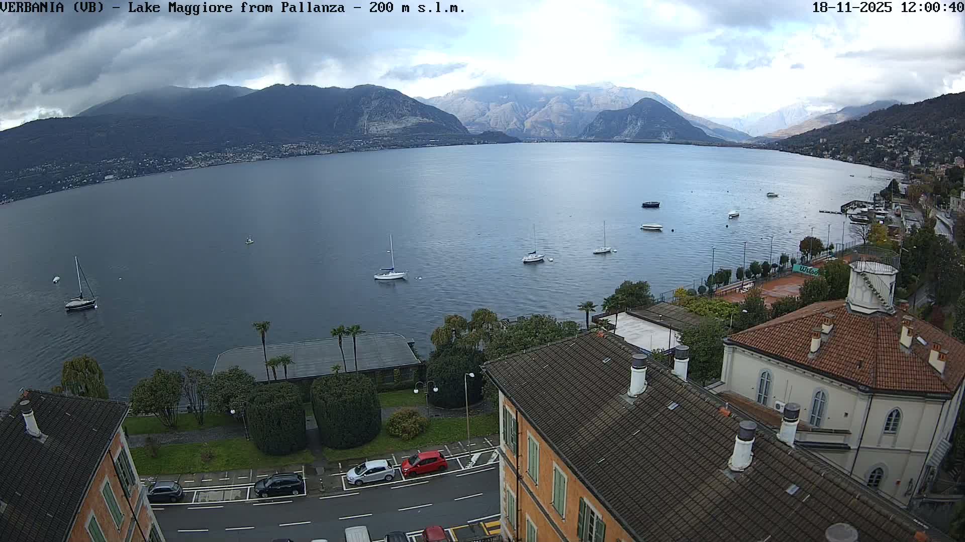An expansive lake, dotted with numerous boats and bordered by residential buildings and green trees along its shores, appears under an overcast and hazy sky, indicating a gloomy, possibly rainy day.