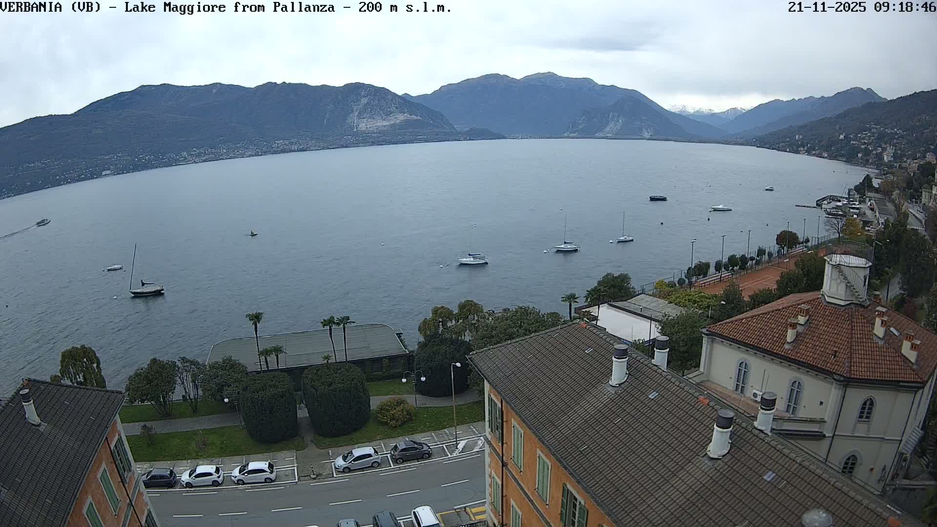 A panoramic view shows a calm lake dotted with boats, surrounded by green and rocky mountains, with a bustling town featuring buildings, parked cars, and a tennis court along the near shore under a cloudy sky.