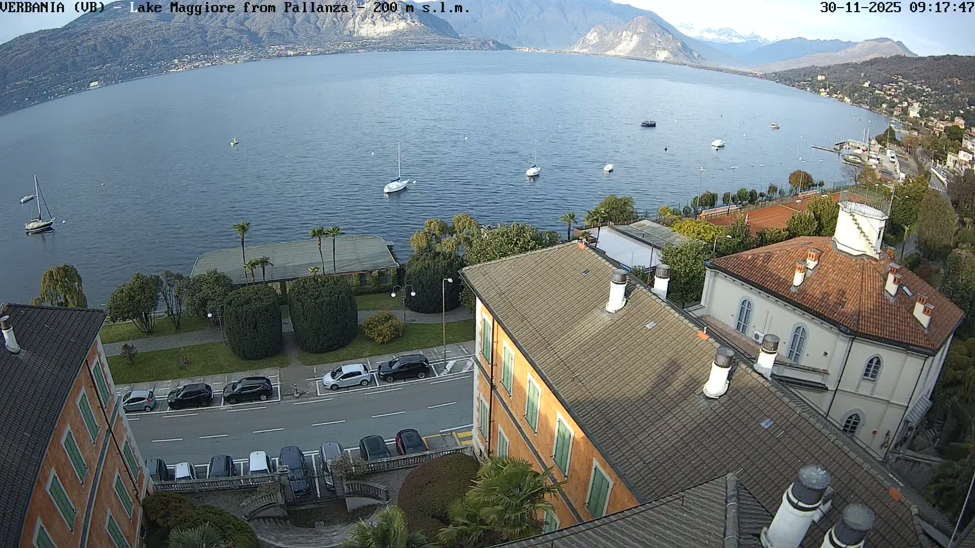 On a clear day, an elevated view captures a town's buildings and a road with parked cars bordering a calm blue lake featuring several boats, with forested and snow-capped mountains in the distant background.