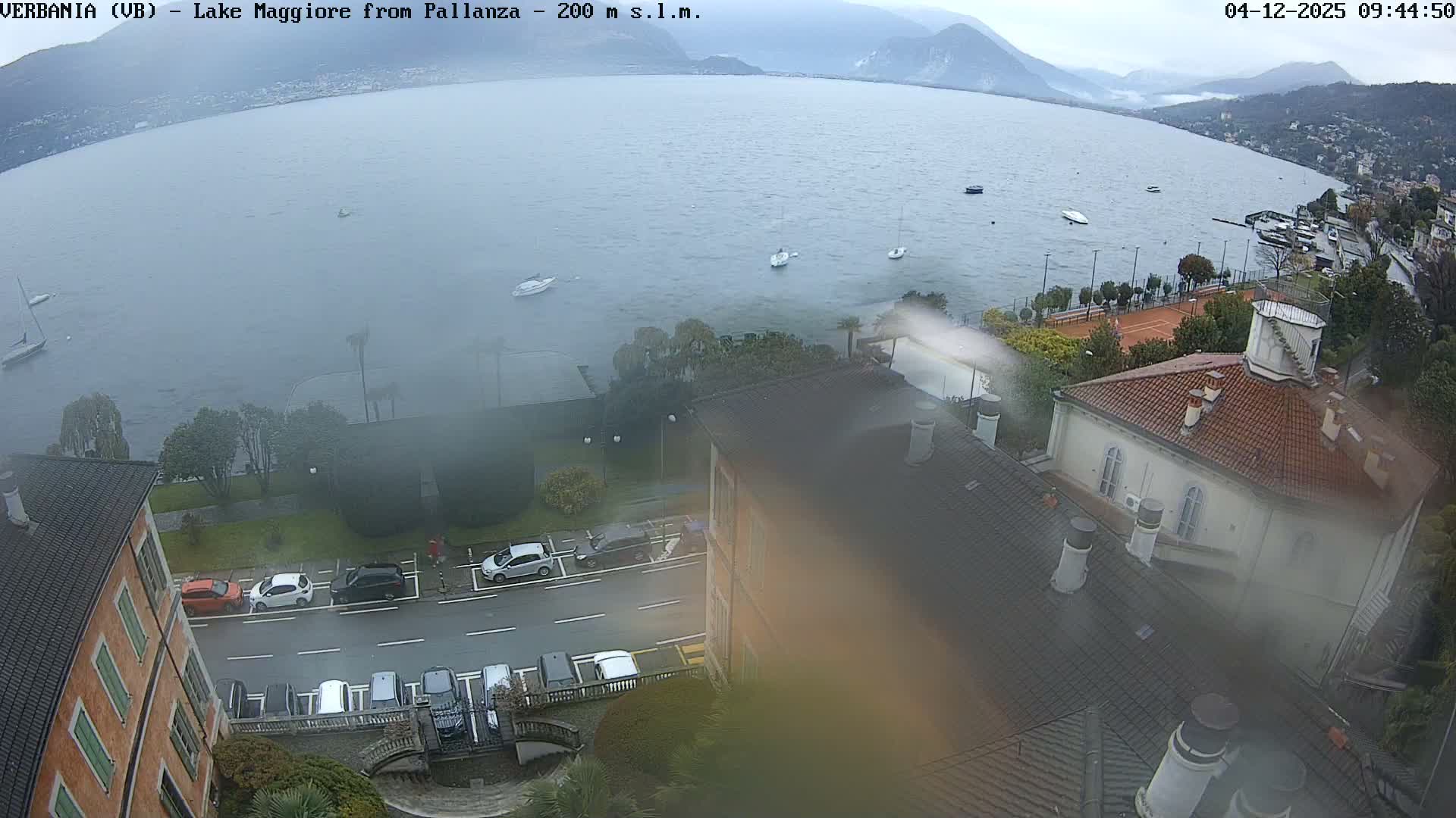 An elevated view captures a wide, grey lake with several boats and hazy mountains in the distance, while the foreground shows a lakeside road with parked cars, buildings with tiled roofs, and some trees under an overcast sky on a dull day.