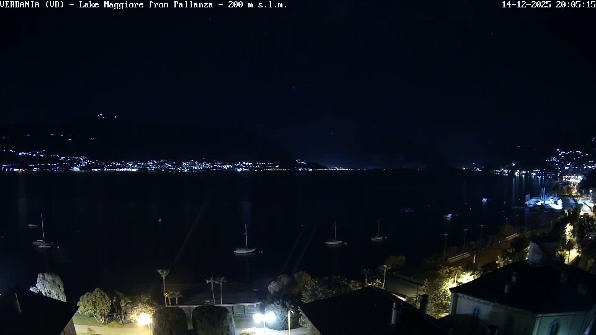 An elevated view captures a wide, grey lake with several boats and hazy mountains in the distance, while the foreground shows a lakeside road with parked cars, buildings with tiled roofs, and some trees under an overcast sky on a dull day.