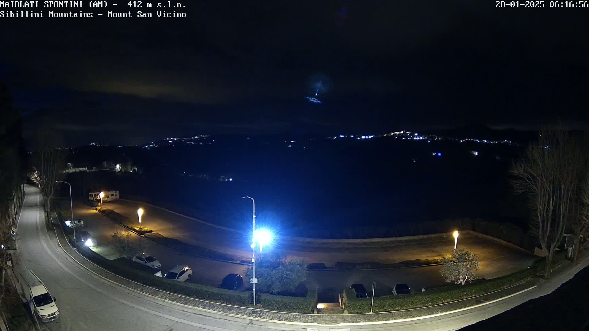 A nighttime view of a parking lot and roadway, illuminated by streetlights, overlooking a distant town with faint lights under a dark, clear sky.