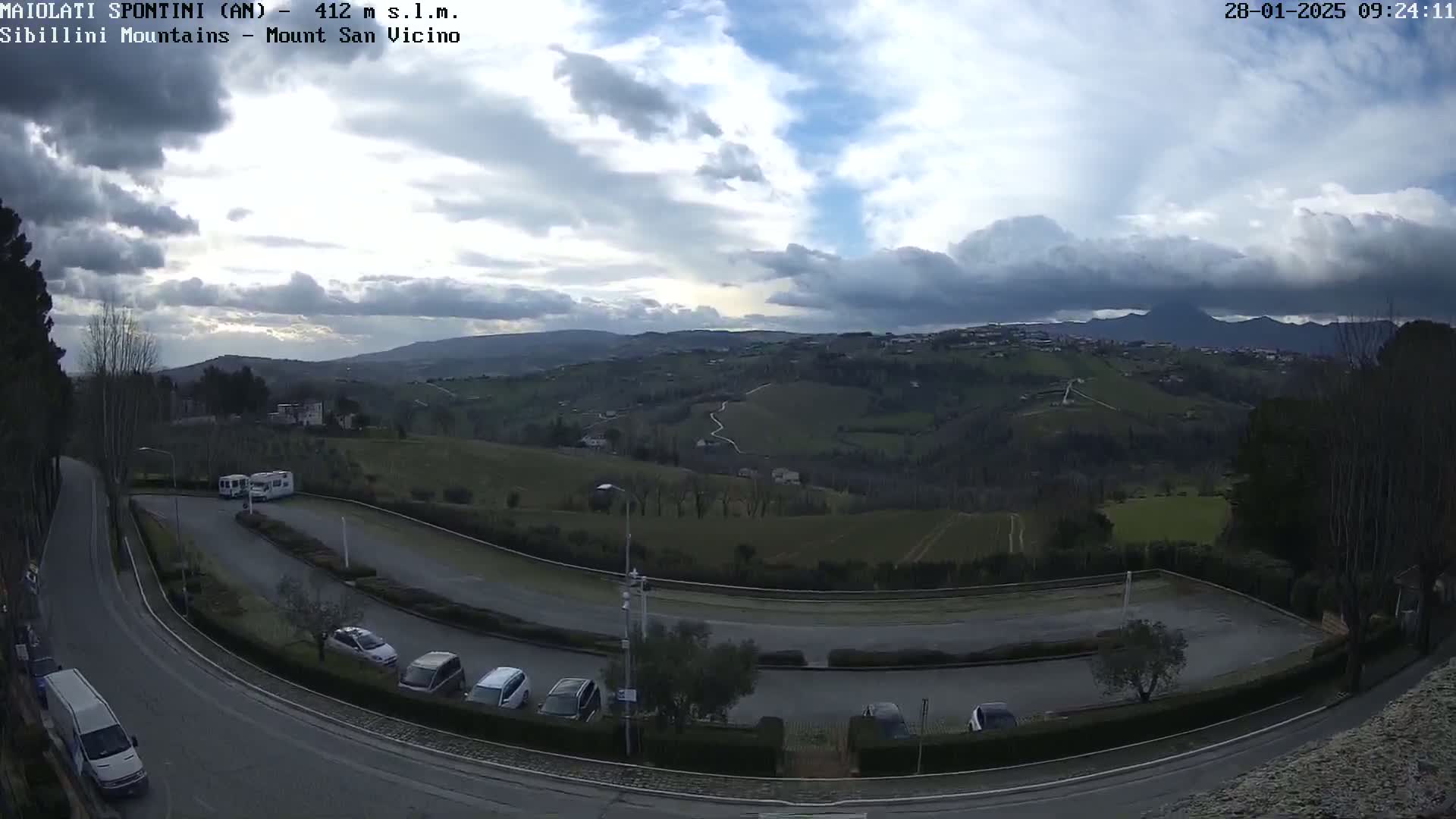 A mostly cloudy sky overlooks a parking lot with several cars and a van, curving roads, and rolling hills dotted with buildings in the distance.