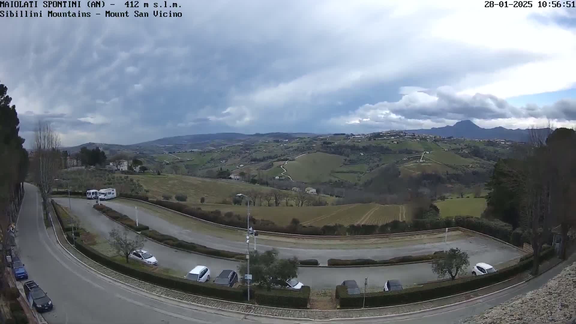 An empty parking lot curves around a hill overlooking a sprawling, gently rolling landscape under a cloudy sky.