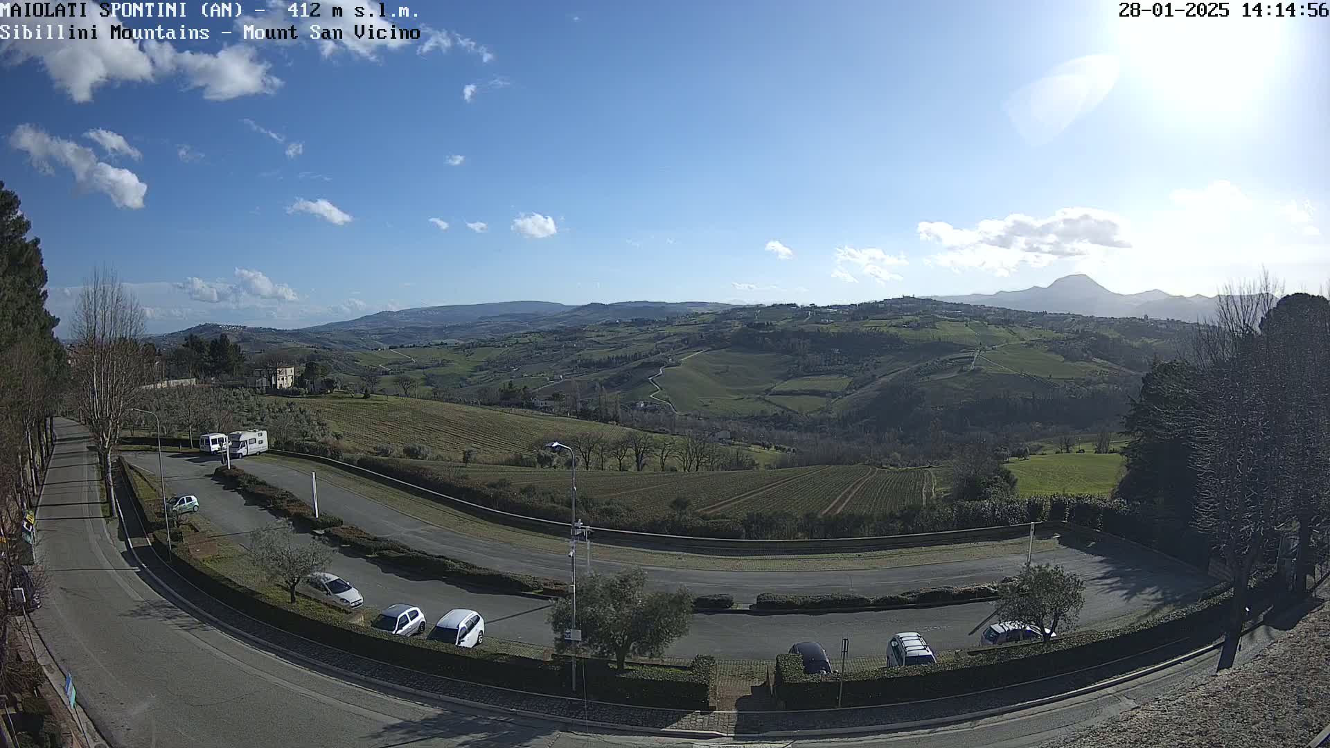 A sunny day reveals a parking lot with several cars, a curving road, and a vast, rolling landscape of green hills and distant mountains under a mostly clear blue sky.