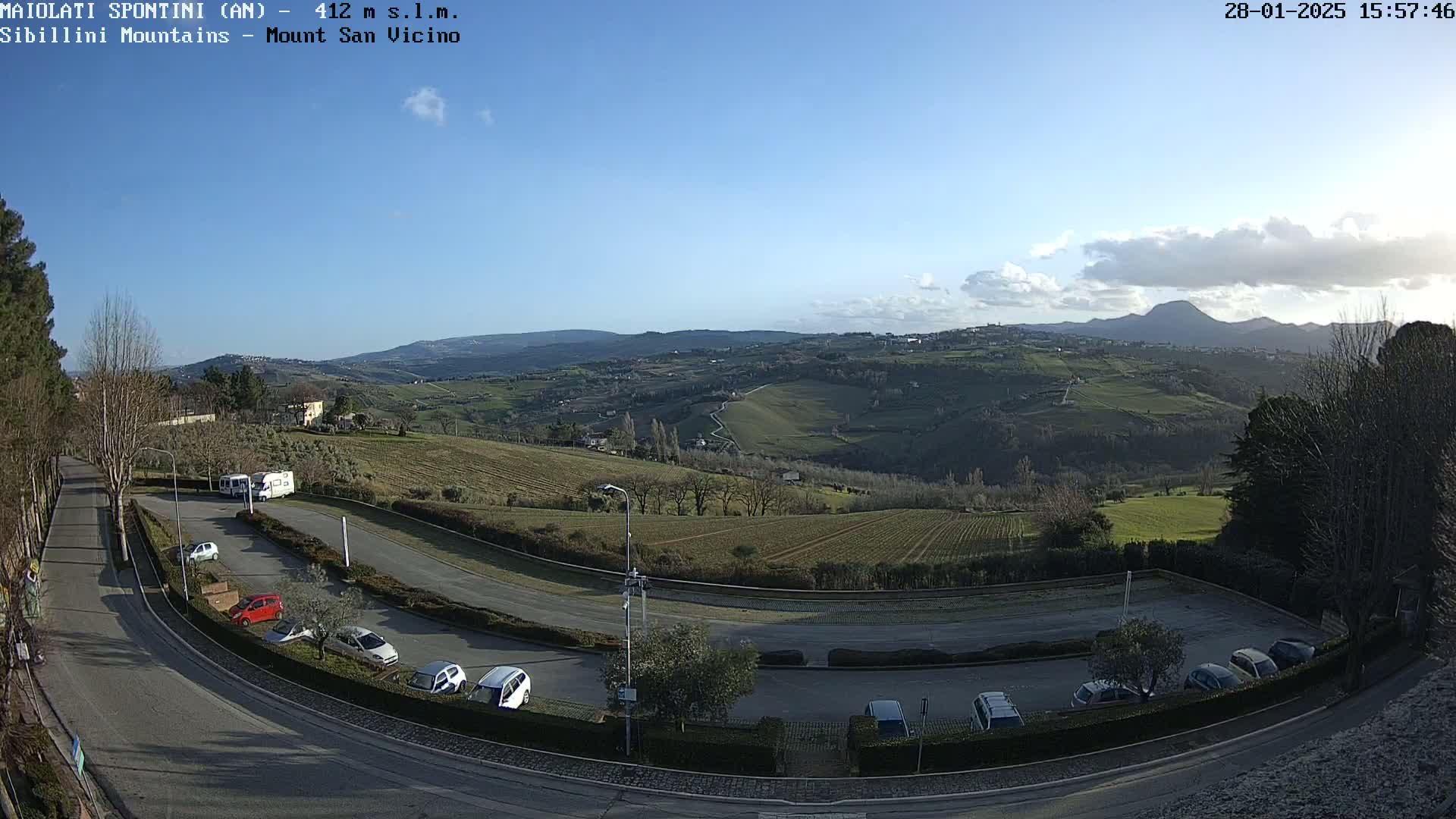 A mostly sunny day reveals a hilly landscape with vineyards and a parking lot containing several cars and camper vans, overlooked by a winding road.