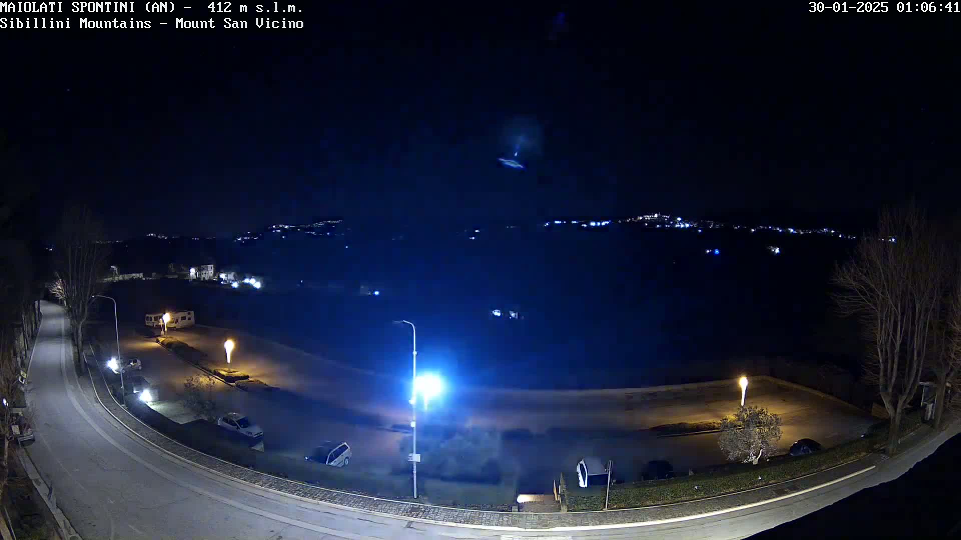 A nighttime, overhead view of a mostly empty parking lot and road, illuminated by streetlights, with a distant town visible in the background under a clear, dark sky.