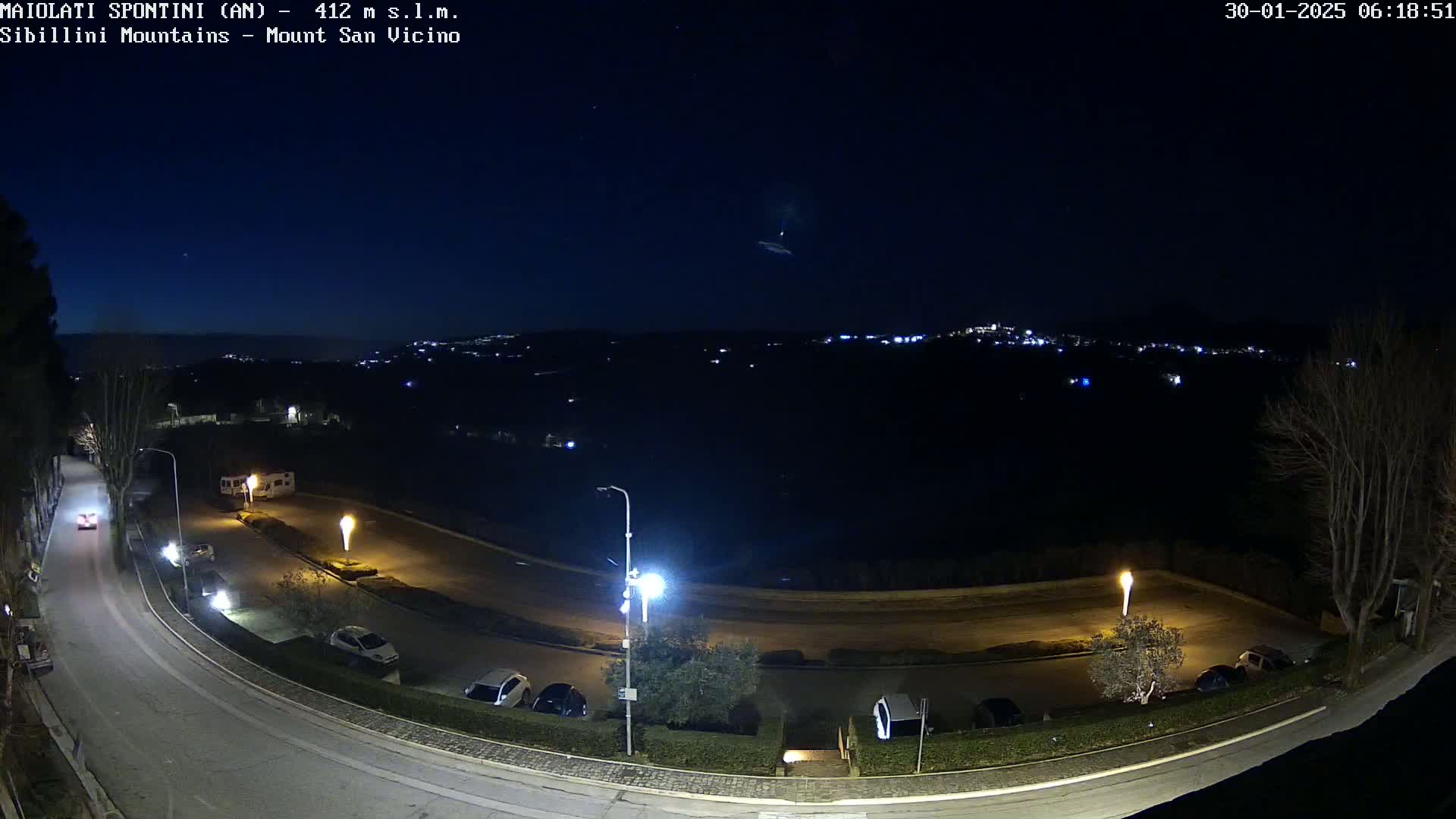 A nighttime, high-angle view shows a curving road with parked cars and a few streetlights, overlooking a dark landscape with distant city lights under a clear, starry sky.