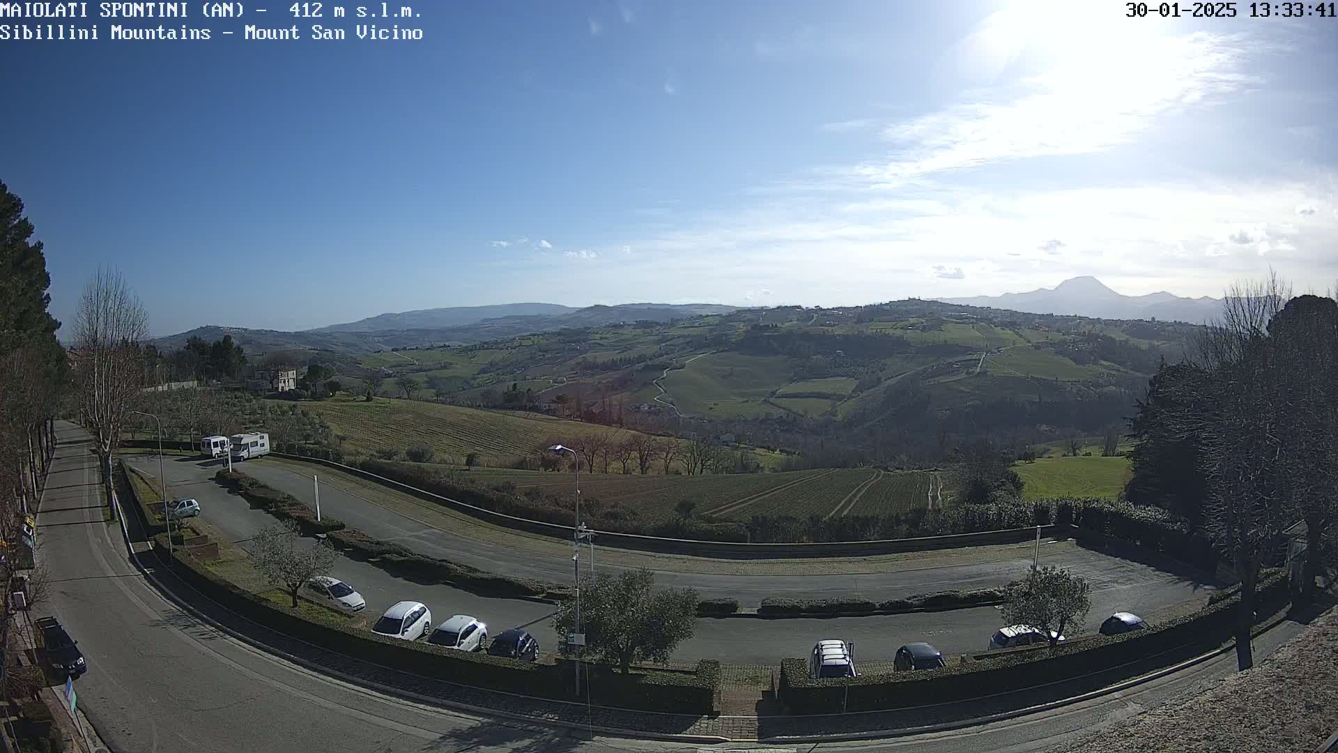 A sunny, clear day overlooks a curved roadway and parking lot nestled among rolling green hills and vineyards, with mountains visible in the distance.