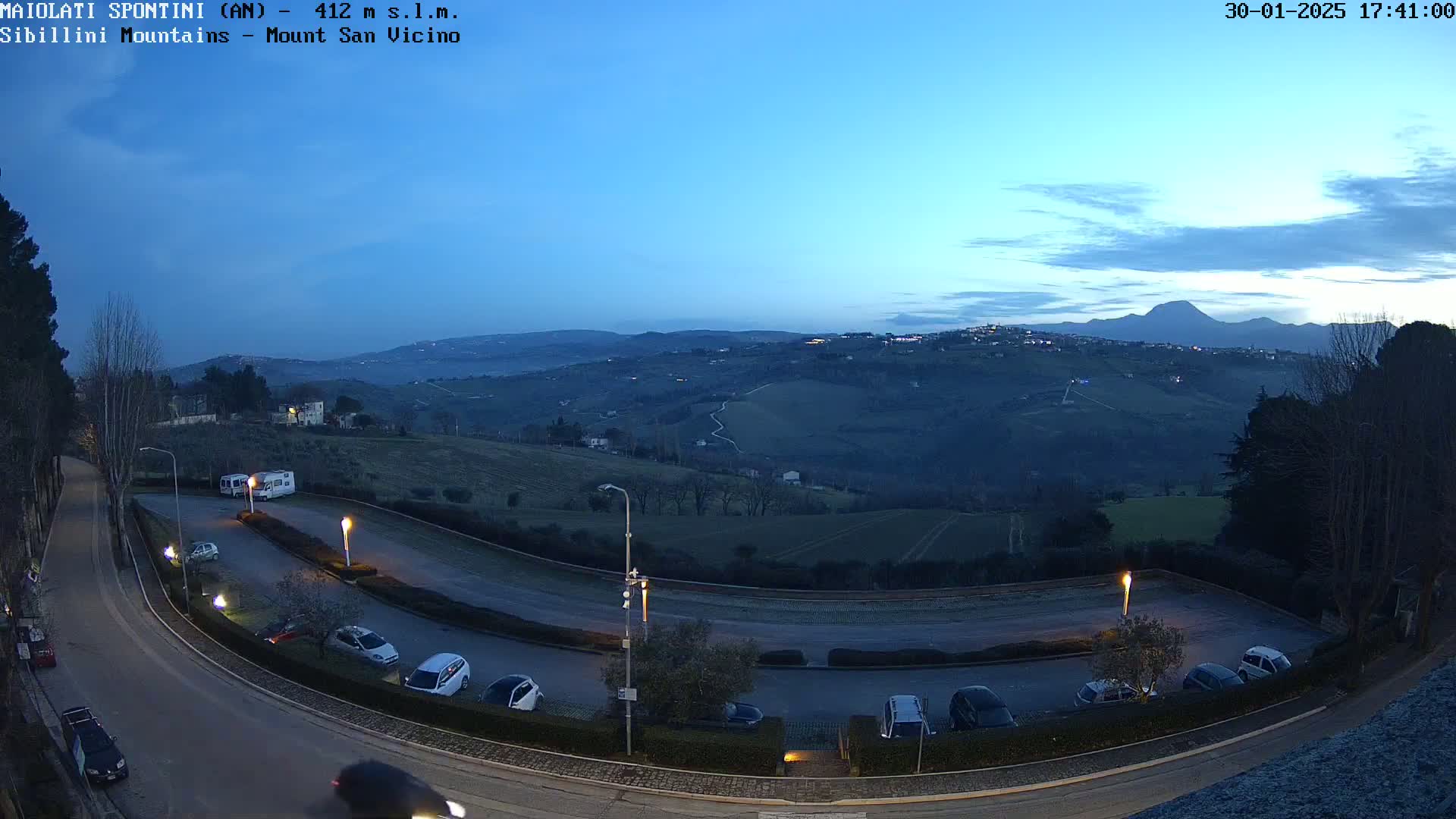 A twilight view of a parking lot curving around a hill, overlooking a valley with distant mountains under a clear, dusky sky.