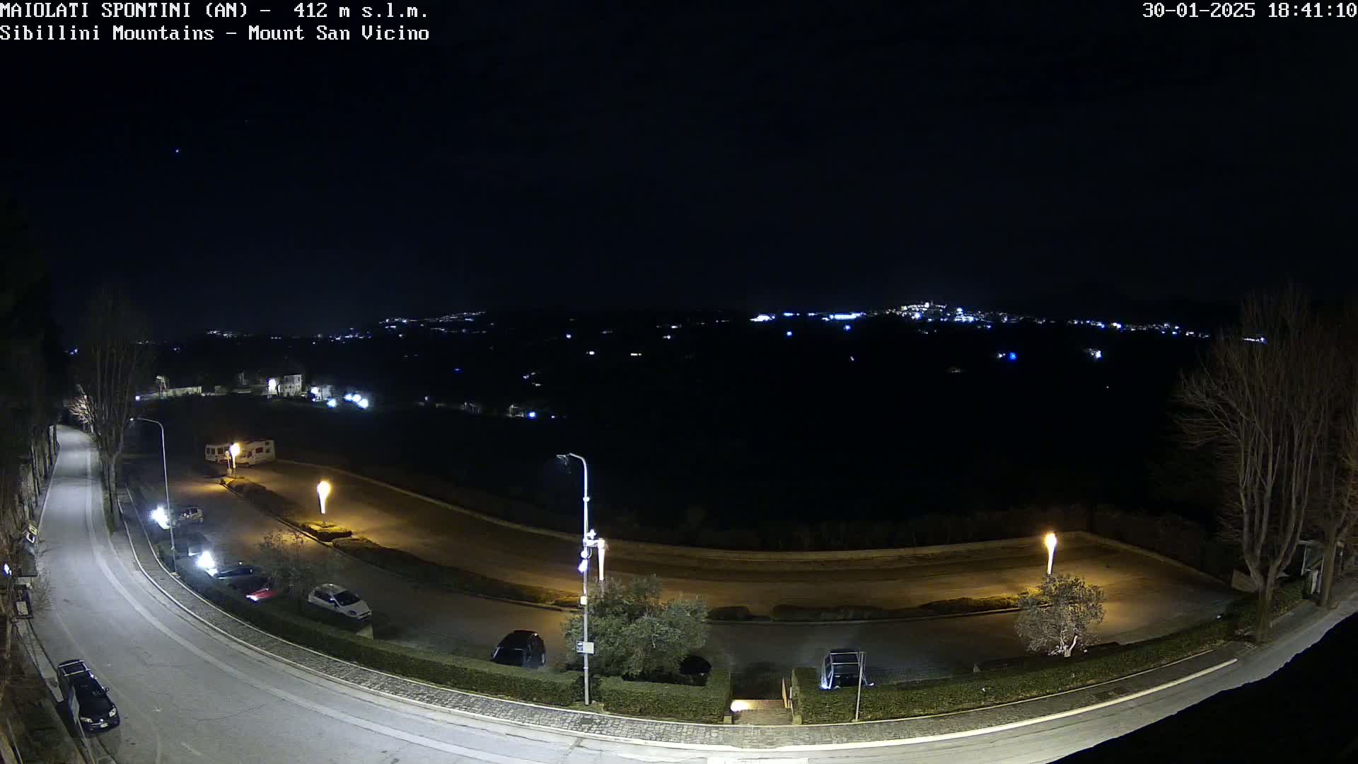 A nighttime, high-angle view of a curved road with parked cars and illuminated lampposts, overlooking a distant town speckled with lights.