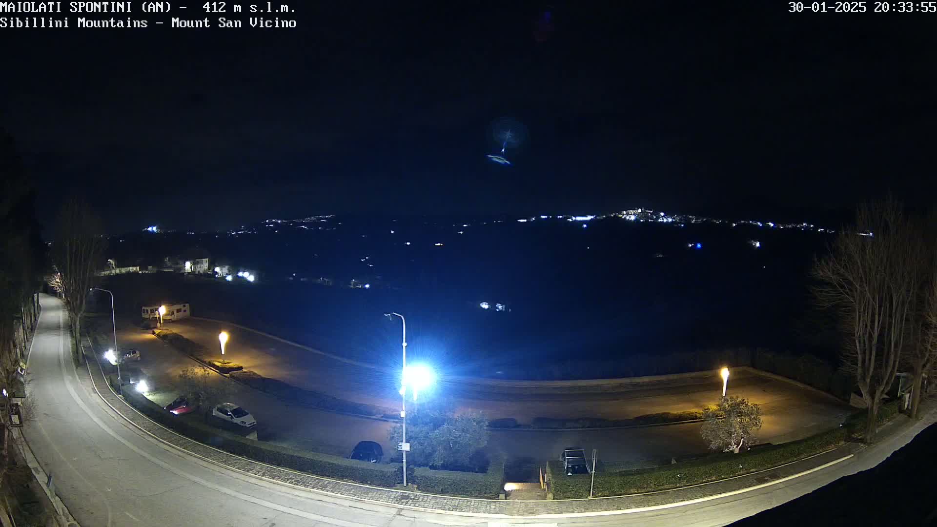 A nighttime, high-angle view of a curving road illuminated by streetlights, a few parked cars, and a distant town twinkling with lights under a dark, clear sky.