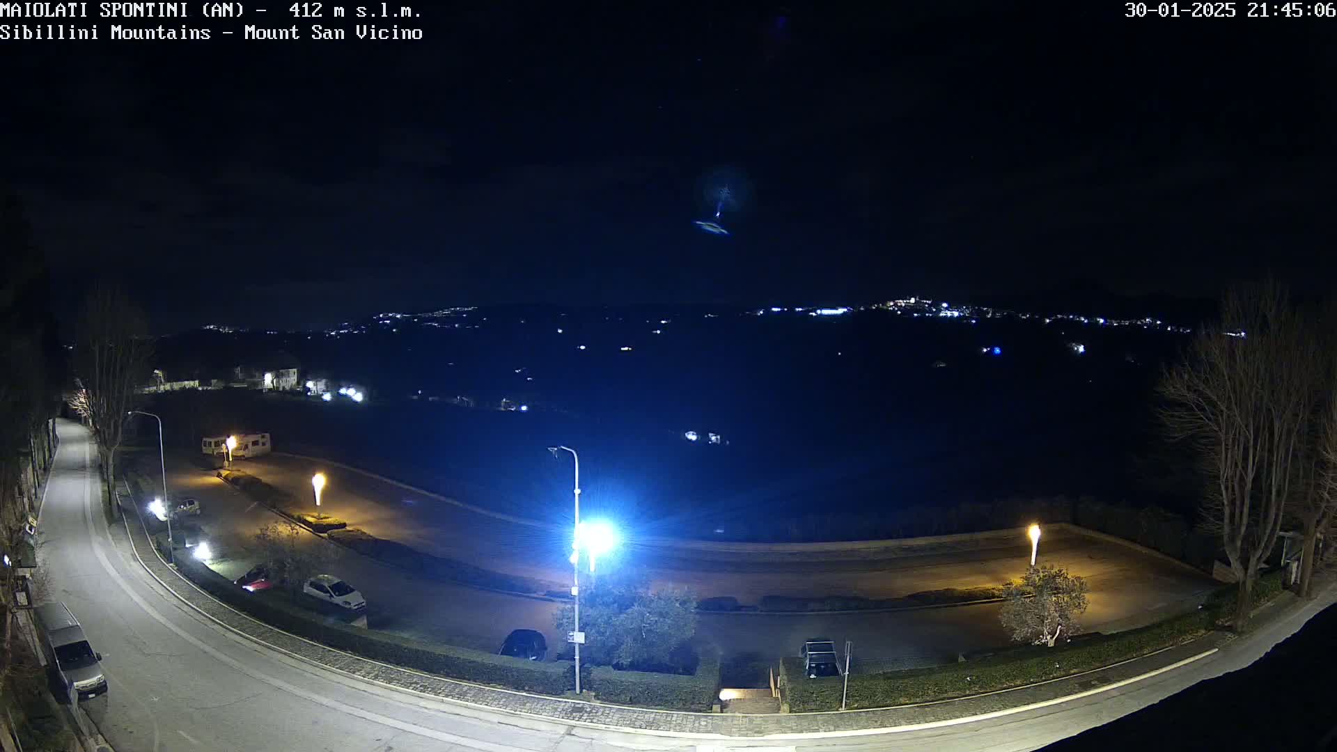 A nighttime view of a curving road and parking area, illuminated by streetlights, overlooks a distant town nestled in dark hills under a clear night sky with a faint light streak.