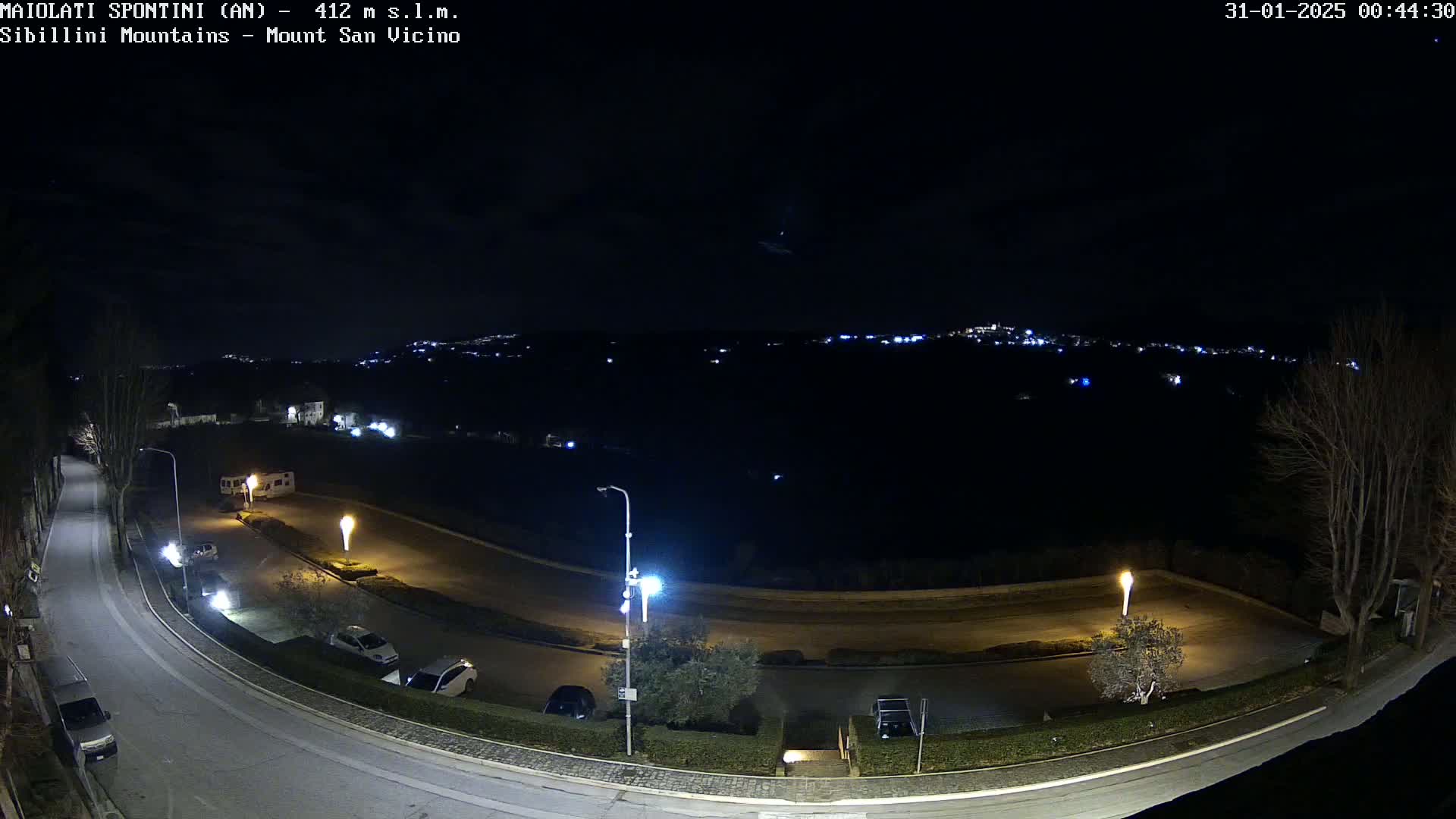 A nighttime, overhead view of a curving road with parked cars and lampposts, leading to a distant, faintly lit town under a clear, dark sky.