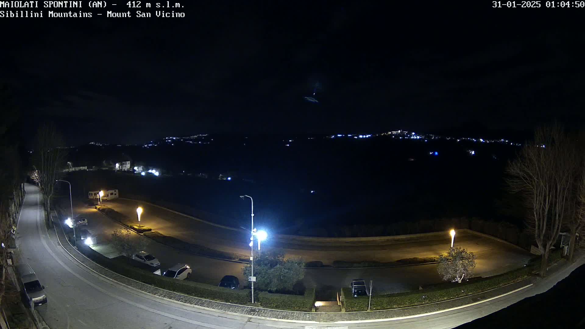 A nighttime, overhead view of a curving road illuminated by streetlights, with parked cars and a distant, dimly lit town visible under a clear, dark sky.