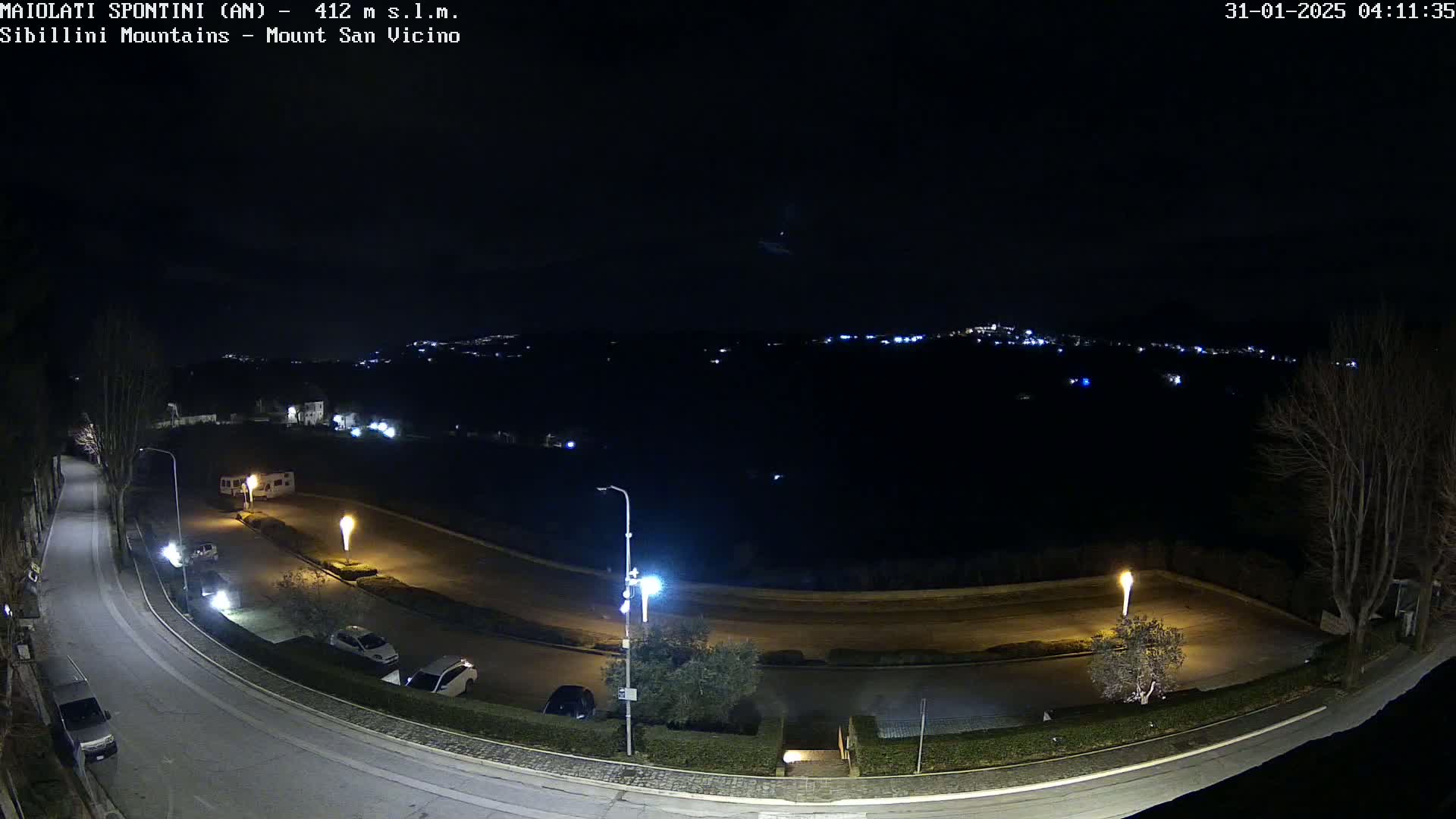 A nighttime, overhead view of a curved road with parked cars and a few illuminated lights, overlooking a dark landscape with distant, faintly visible lights.