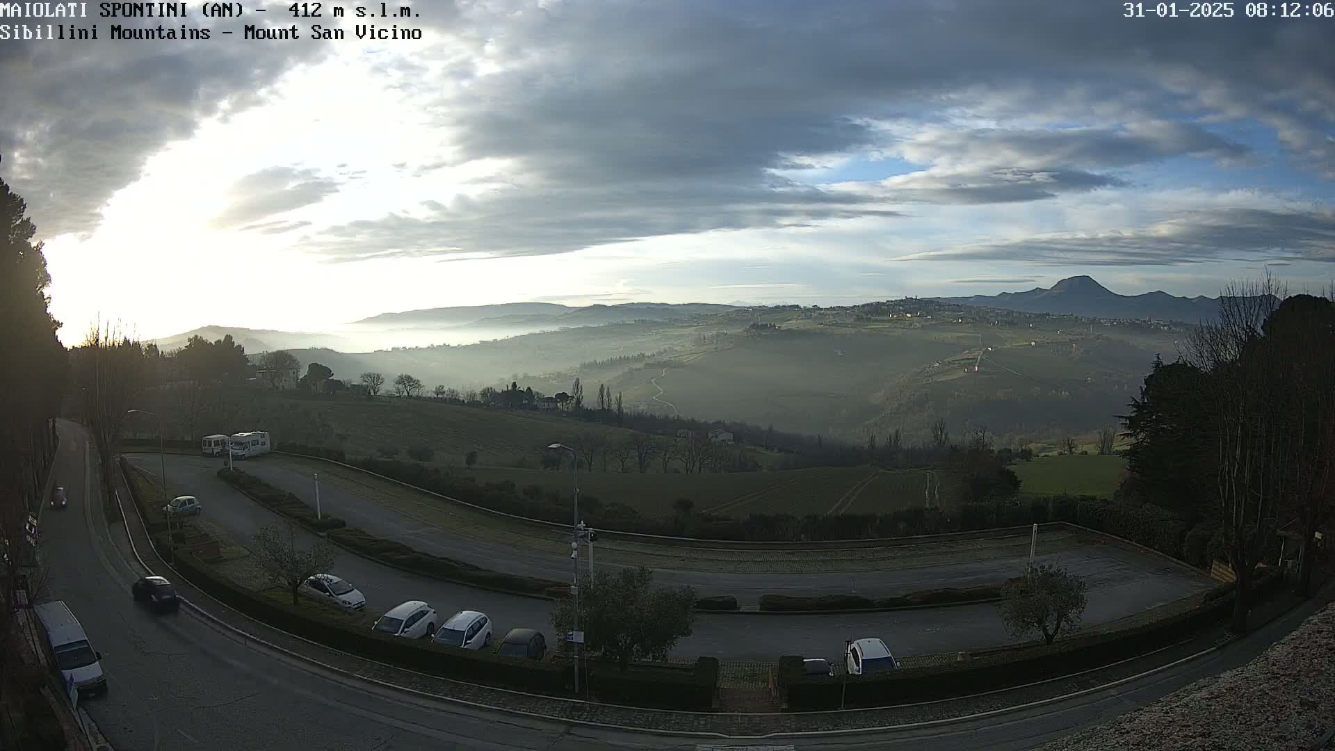 A hazy sunrise reveals a hilly landscape with a parking lot and road in the foreground, under a partly cloudy sky.
