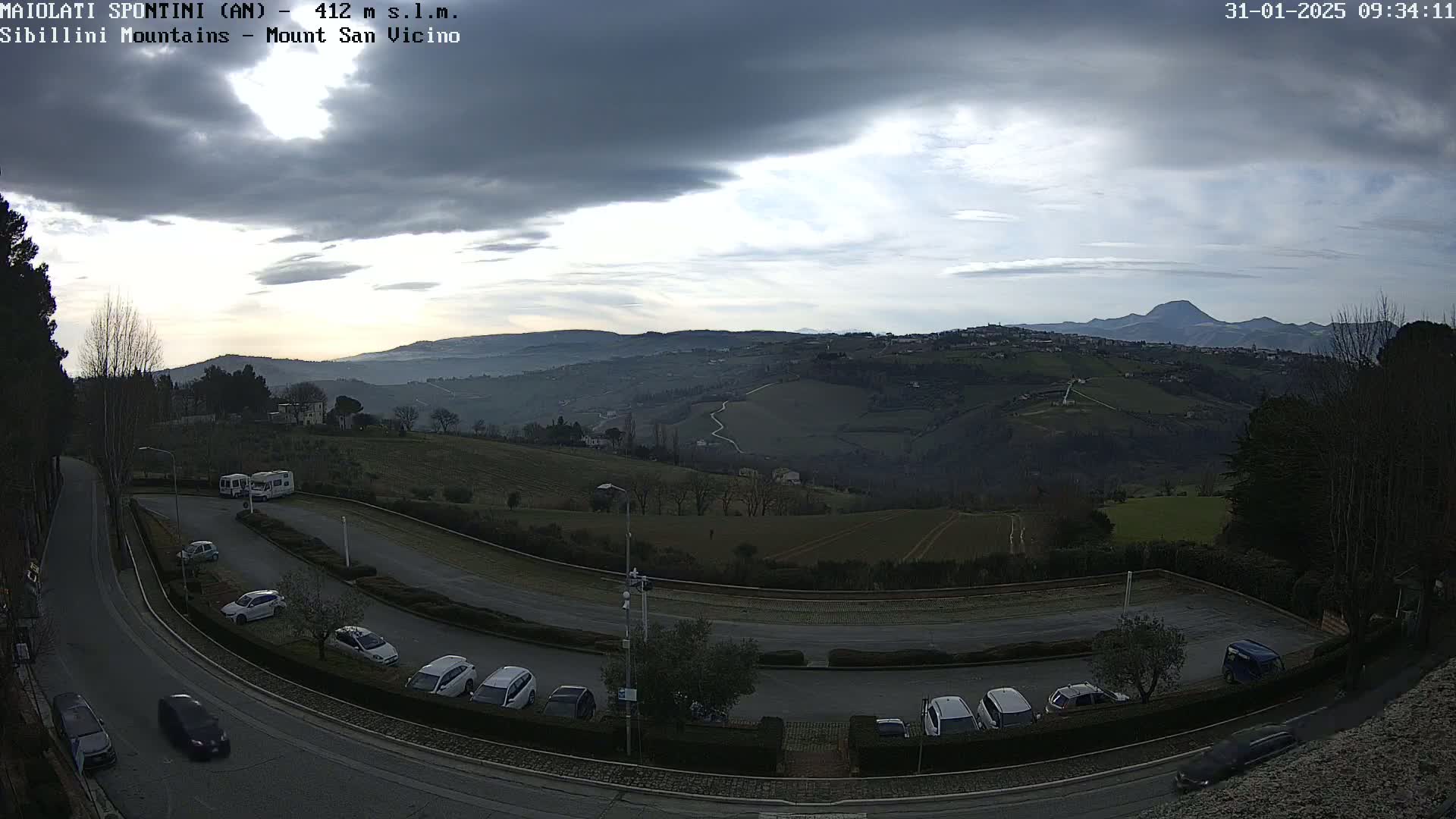 A partly cloudy, overcast sky overlooks a hilly landscape with a parking lot containing several cars and a few RVs, and a winding road leading toward distant mountains.