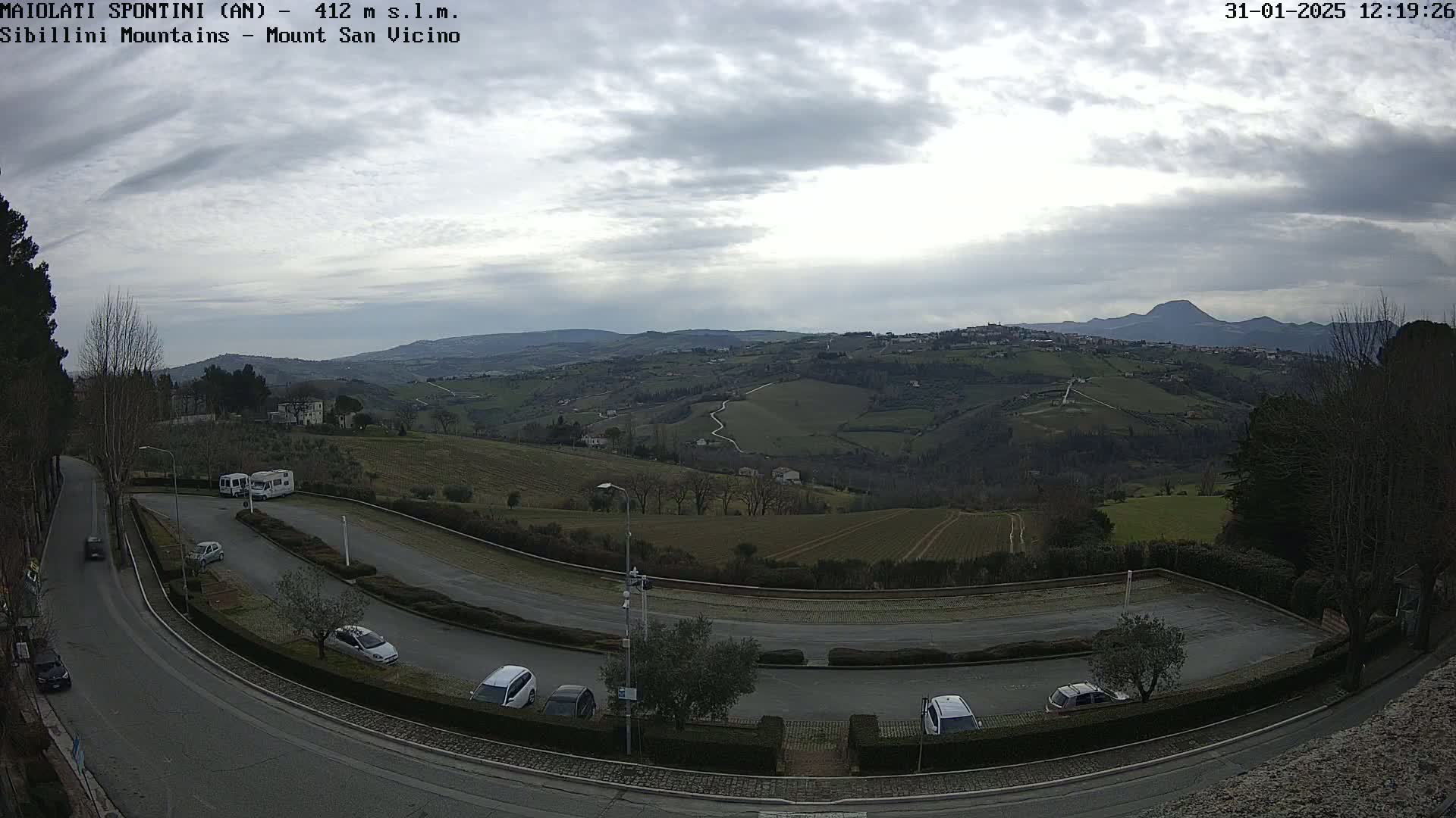 A paved parking lot and curved road are visible with several cars parked, overlooking a hilly landscape under a cloudy sky.
