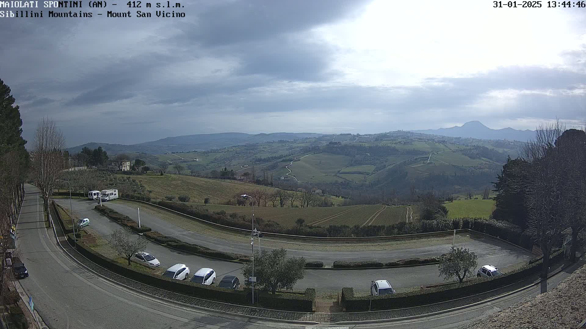 A parking lot with several cars and camper vans overlooks a hilly landscape under a partly cloudy sky.
