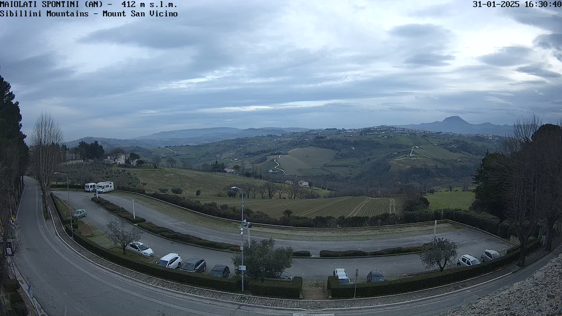A mostly cloudy sky overlooks a paved parking lot with several cars and camper vans situated on a hilltop overlooking a valley of rolling green hills and distant mountains.