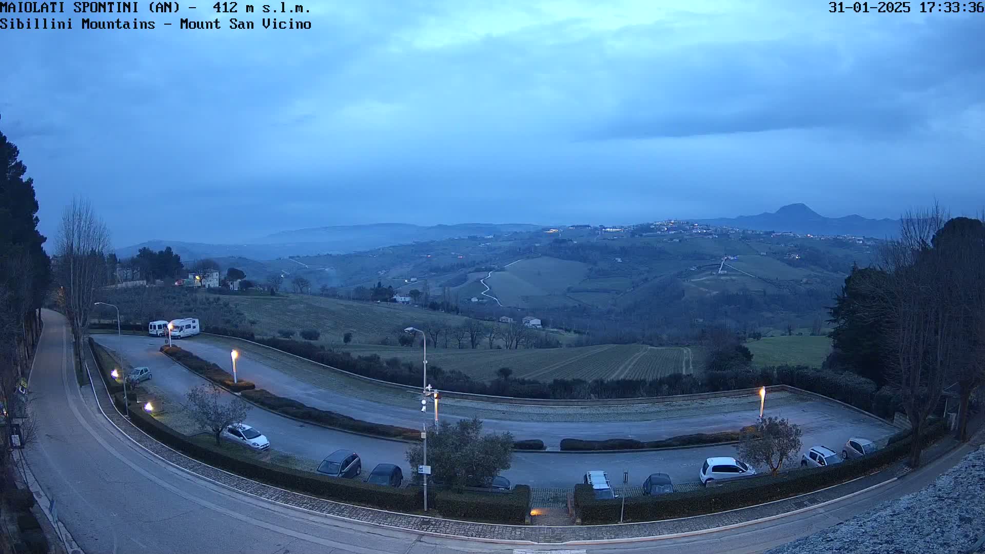 A curved road with parked cars overlooks a hilly landscape under a twilight sky.