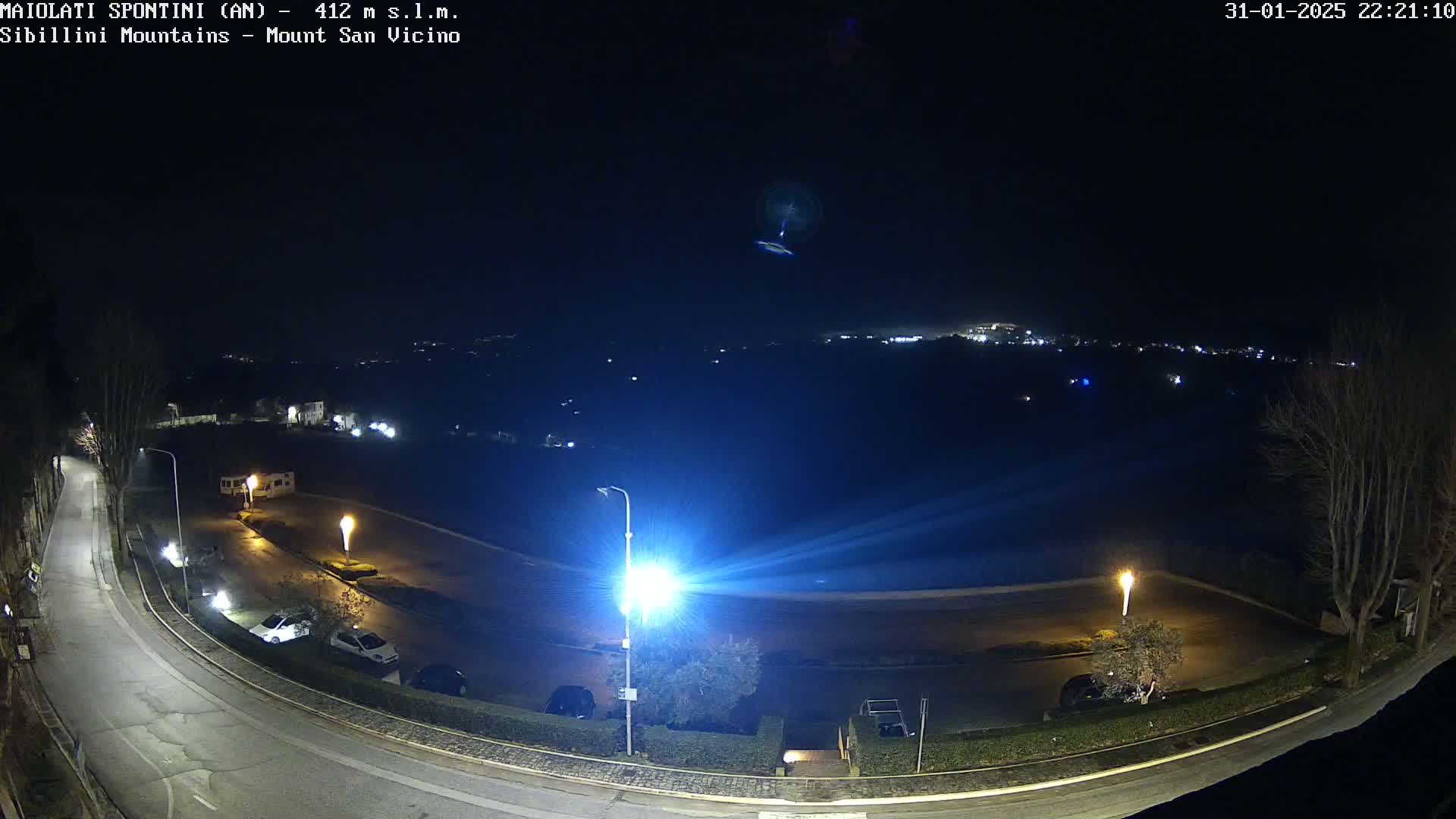 A nighttime, high-angle view of a parking lot and curving road illuminated by bright lights, overlooking a dimly lit town nestled in the distance under a clear, dark sky.