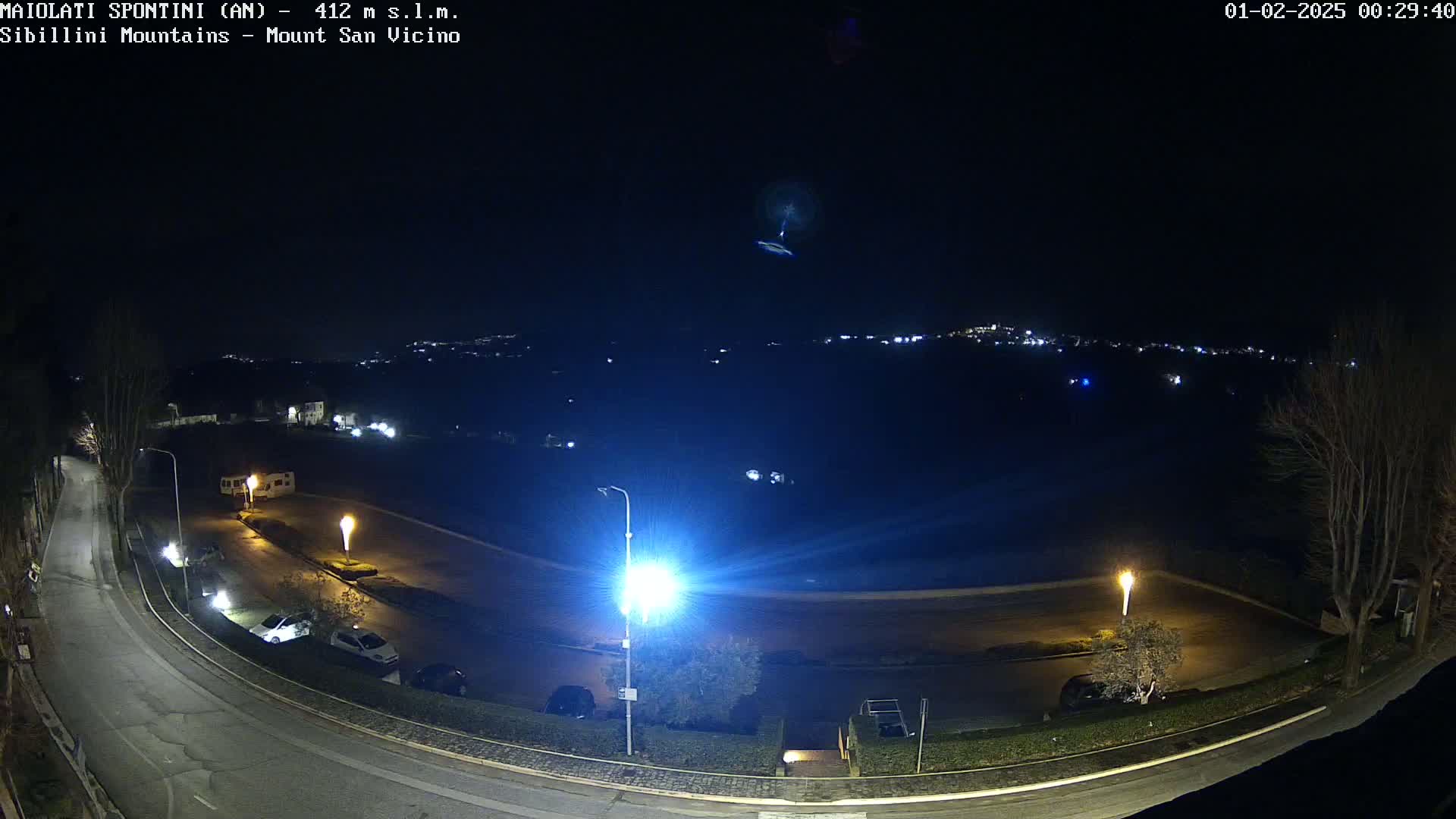 A nighttime, overhead view of a mostly empty parking lot and roadway, with a distant town illuminated in the background under a clear, dark sky.