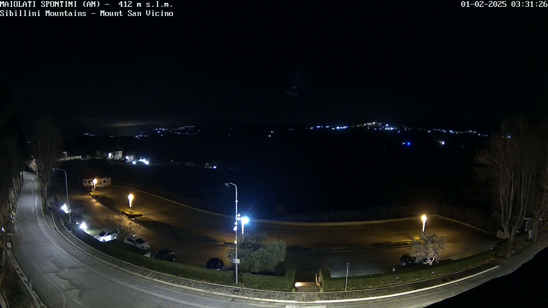 A nighttime, high-angle view shows a curved road with parked cars and streetlights, overlooking a dark landscape with distant, faintly visible lights.