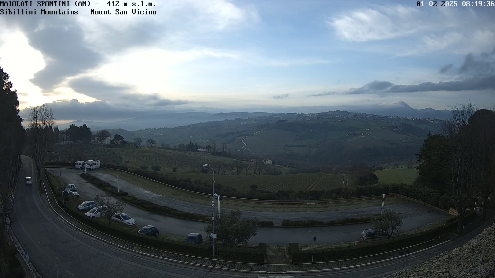 A wide shot shows a parking lot and winding road overlooking a hilly landscape under a mostly cloudy sky.